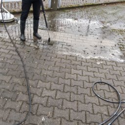 Michał Nowak - Czyszczenie kostki brukowej myjką ciśnieniową. Widoczny fragment kostki przed i po czyszczeniu. W tle ogrodzenie.