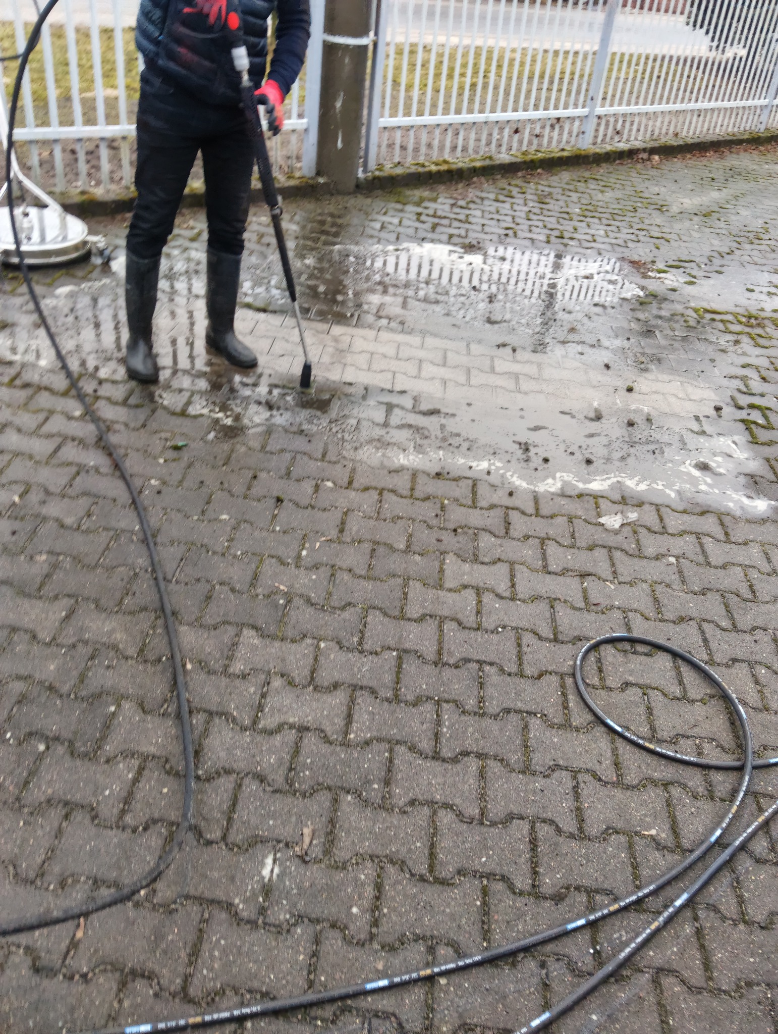 Czyszczenie kostki brukowej myjką ciśnieniową. Widoczny fragment kostki przed i po czyszczeniu. W tle ogrodzenie.