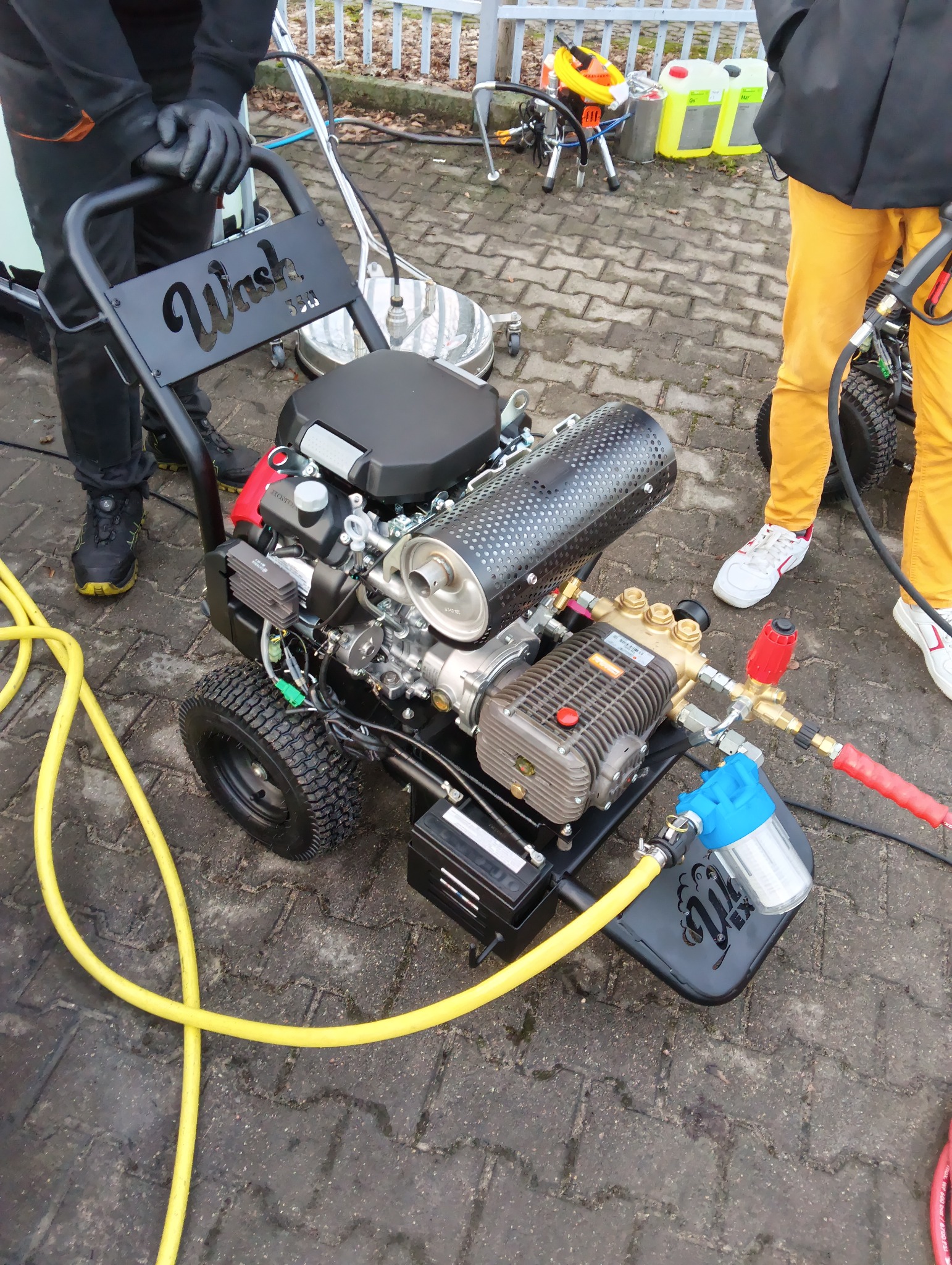 Profesjonalna, mobilna myjka ciśnieniowa na kostce brukowej. Żółty wąż, silnik spalinowy, filtr wody. Widoczne fragmenty osób obsługujących urządzenie.