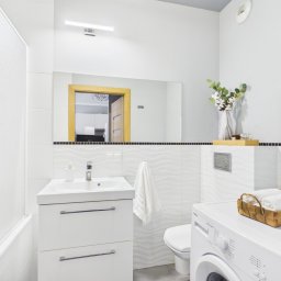 Evesta Sp. z o.o. - Jasna łazienka z białą armaturą, pralką i dekoracjami. W lustrze odbicie fragmentu kuchni. Przytulne wnętrze, przygotowane do sprzedaży lub wynajmu.