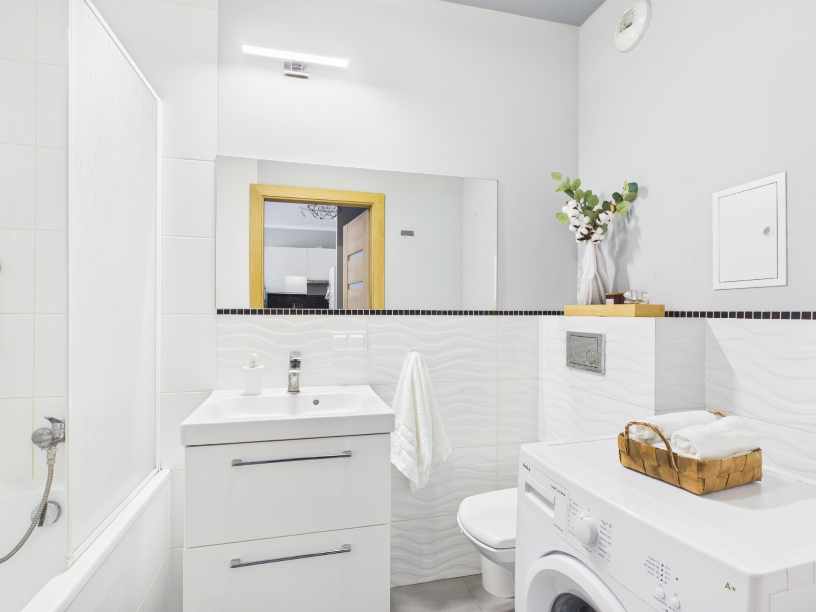 Jasna łazienka z białą armaturą, pralką i dekoracjami. W lustrze odbicie fragmentu kuchni. Przytulne wnętrze, przygotowane do sprzedaży lub wynajmu.