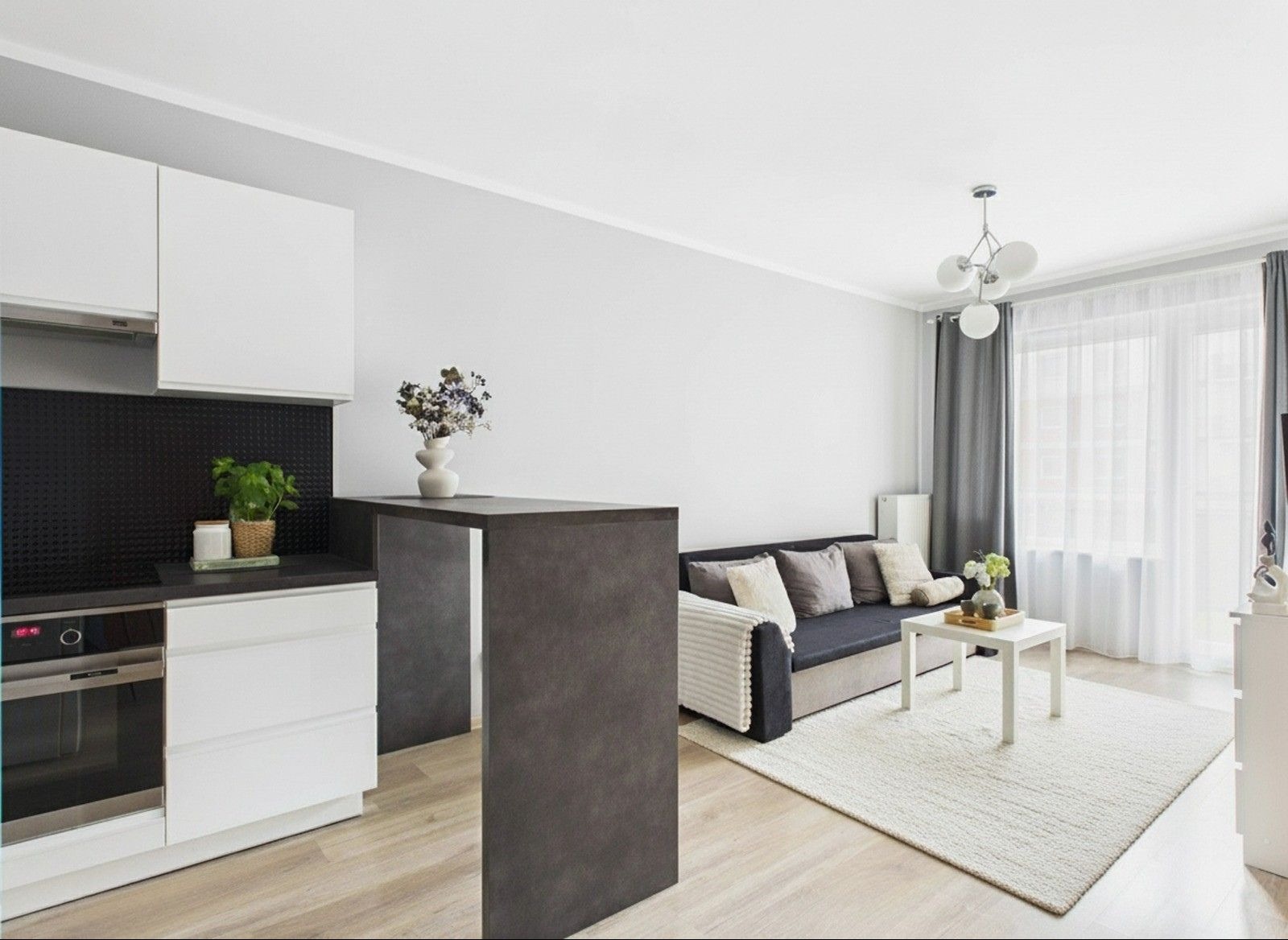 Jasne wnętrze po home stagingu: salon z aneksem kuchennym. Szara sofa z poduszkami, biały dywan, stół. Kuchnia z białymi szafkami i czarnym panelem. Kwiaty w wazonie.