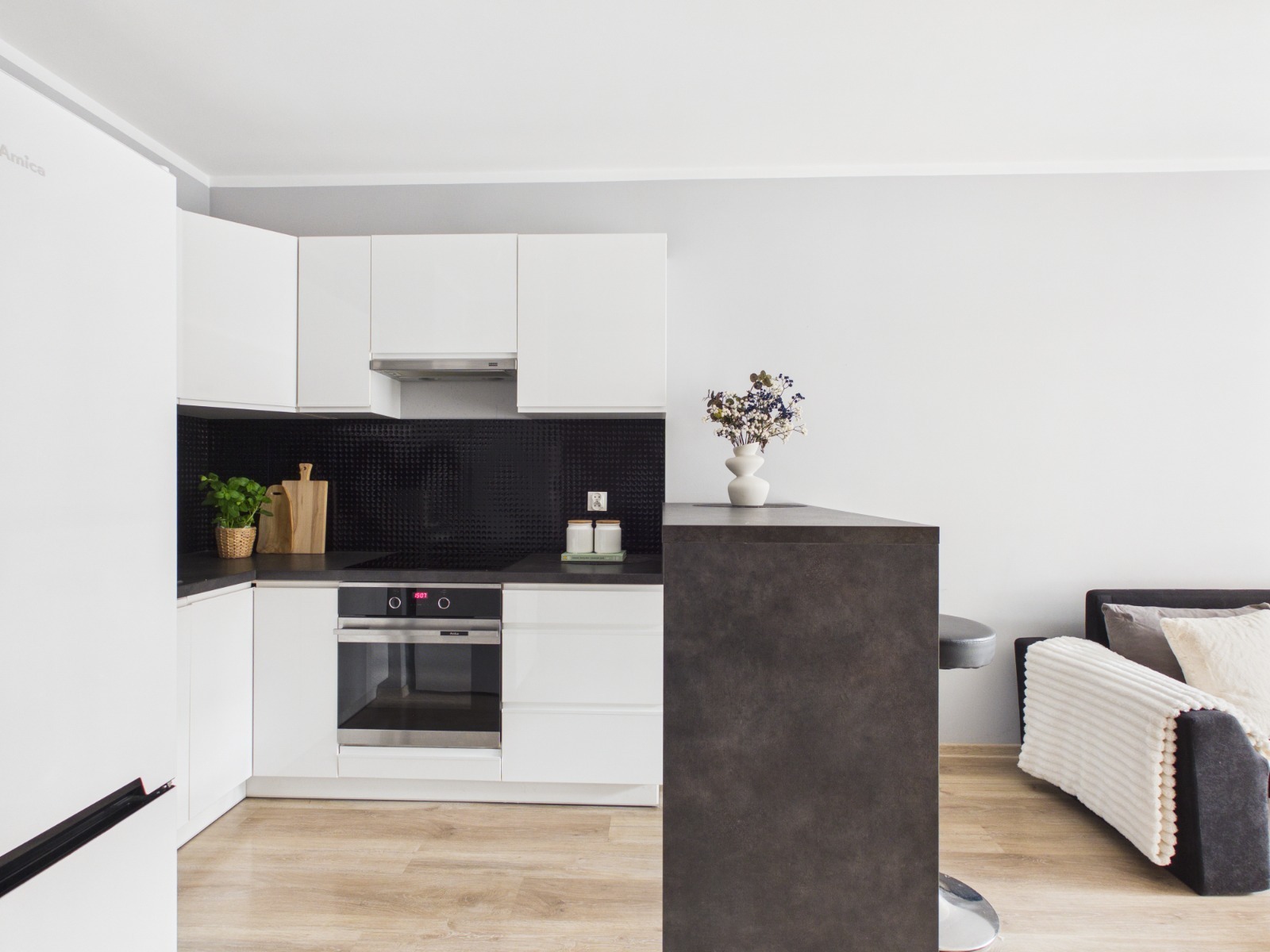 Jasna kuchnia z czarnym panelem, drewnianą deską i kwiatami w wazonie. Minimalistyczny design, nowoczesne oświetlenie, parkiet. Widok na salon z kanapą.