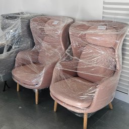 PRZEDSIĘBIORSTWO HANDLOWO-USŁUGOWE MAŁGORZATA RUDNIK - Fotele w folii stretch, gotowe do transportu. Dwa różowe i jeden szary, zabezpieczone przed uszkodzeniami. W tle biała ściana i podłoga z szarych płytek.