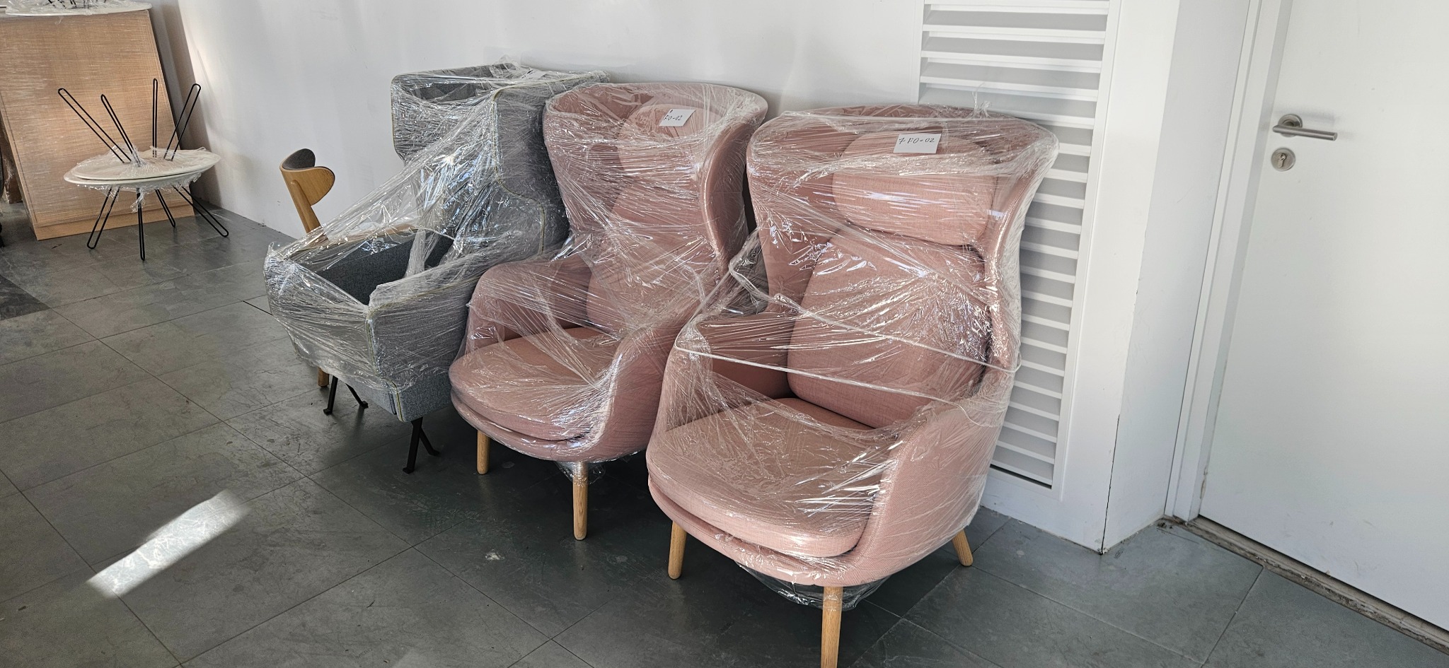 Fotele w folii stretch, gotowe do transportu. Dwa różowe i jeden szary, zabezpieczone przed uszkodzeniami. W tle biała ściana i podłoga z szarych płytek.