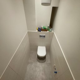 Usługi wykończeniowe Radoń - Wąska, wykafelkowana toaleta w odcieniach szarości i beżu. Podwieszana muszla klozetowa, zabudowa geberit, widoczne akcesoria montażowe. Minimalistyczny design.