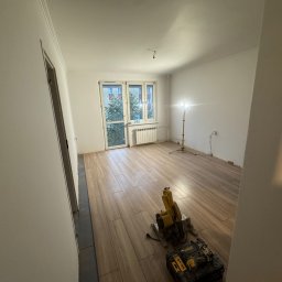 Usługi wykończeniowe Radoń - Układanie paneli podłogowych imitujących drewno w jasnym pokoju. Widoczna piła do cięcia paneli i lampa oświetlająca miejsce pracy. Częściowo wykończone pomieszczenie.