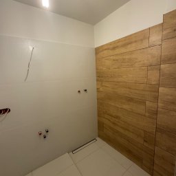 Usługi wykończeniowe Radoń - Wykończona łazienka z drewnopodobnymi i białymi płytkami, widoczne przyłącza wodne i odpływ liniowy. Oświetlenie sufitowe.