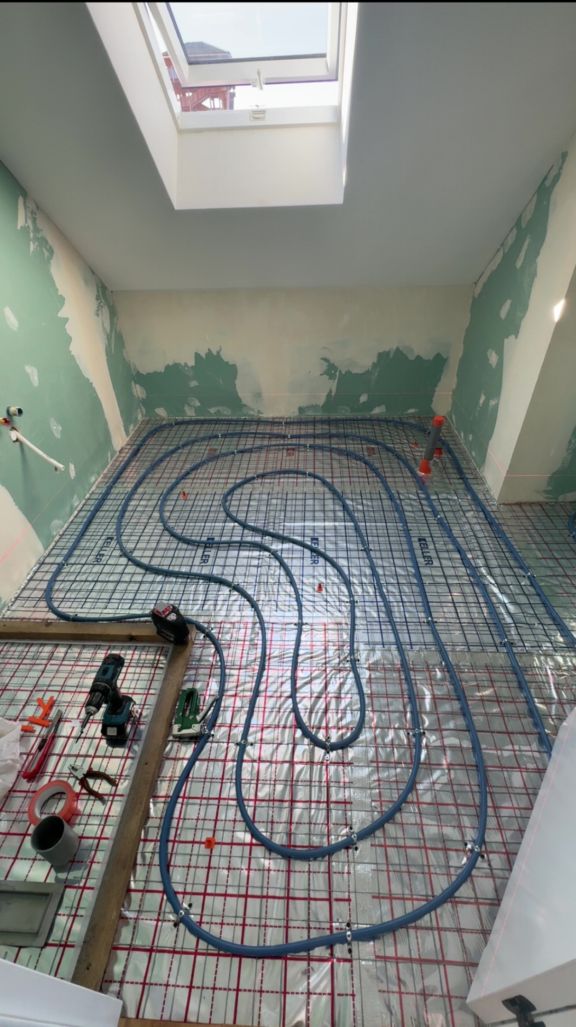Łazienka w trakcie remontu: ułożone rury ogrzewania podłogowego na folii z siatką, narzędzia pracy obok, ściany przygotowane do dalszych prac wykończeniowych, okno dachowe.