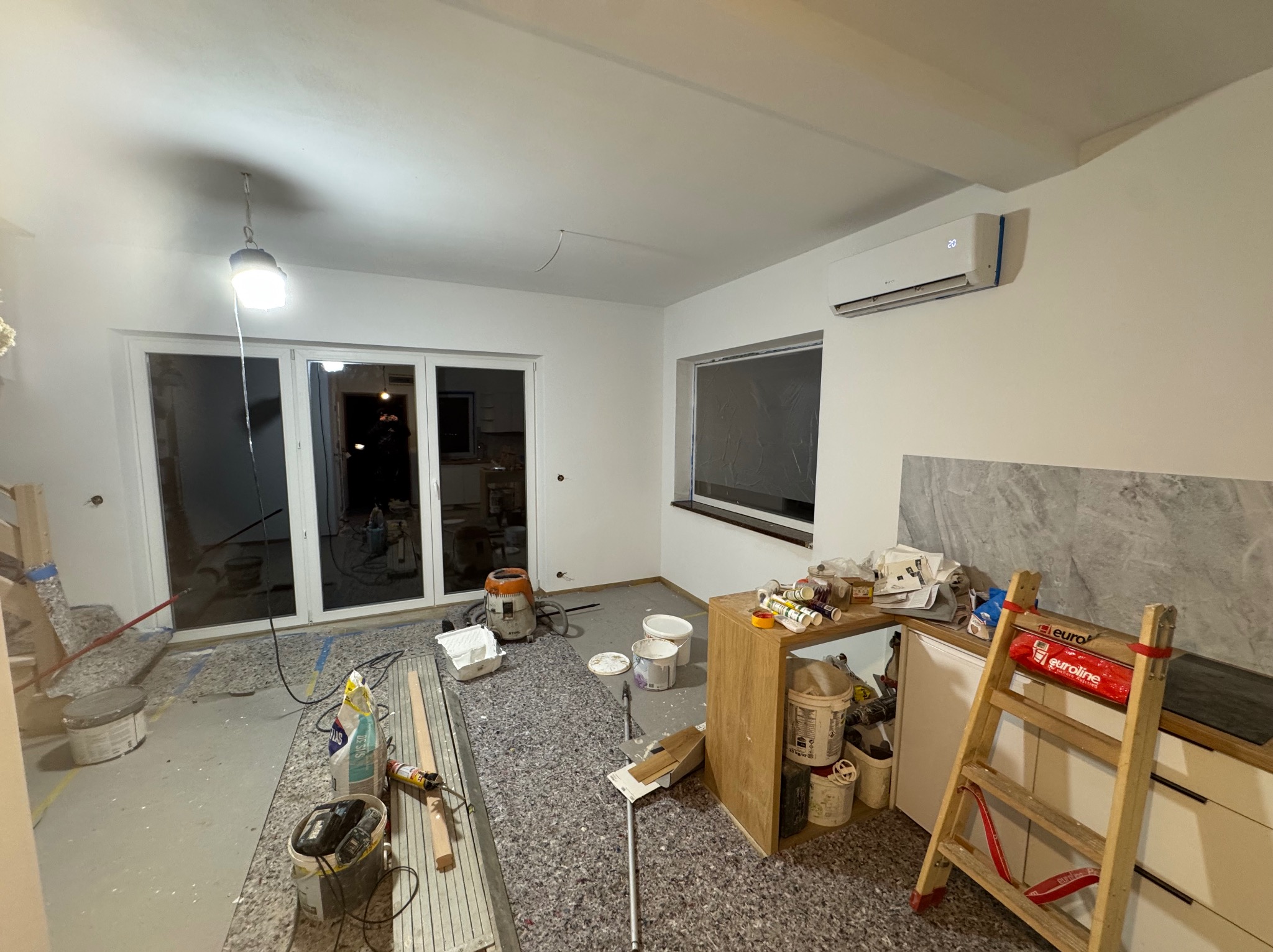 Wnętrze w trakcie remontu: białe ściany, zabezpieczona podłoga, drabina, narzędzia malarskie i wiadro z farbą. Widoczny klimatyzator i okno. Oświetlenie robocze.