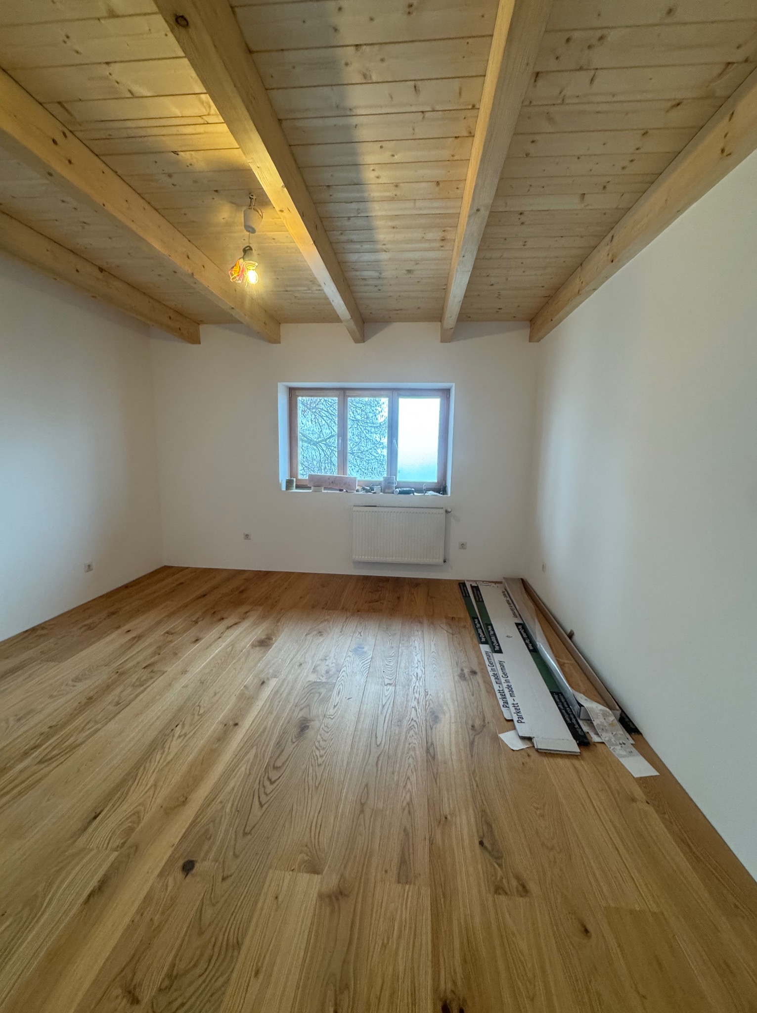 Jasne, drewniane panele podłogowe w pokoju z białymi ścianami i drewnianym sufitem. Przy ścianie leżą paczki paneli, co sugeruje świeżo zakończony montaż.