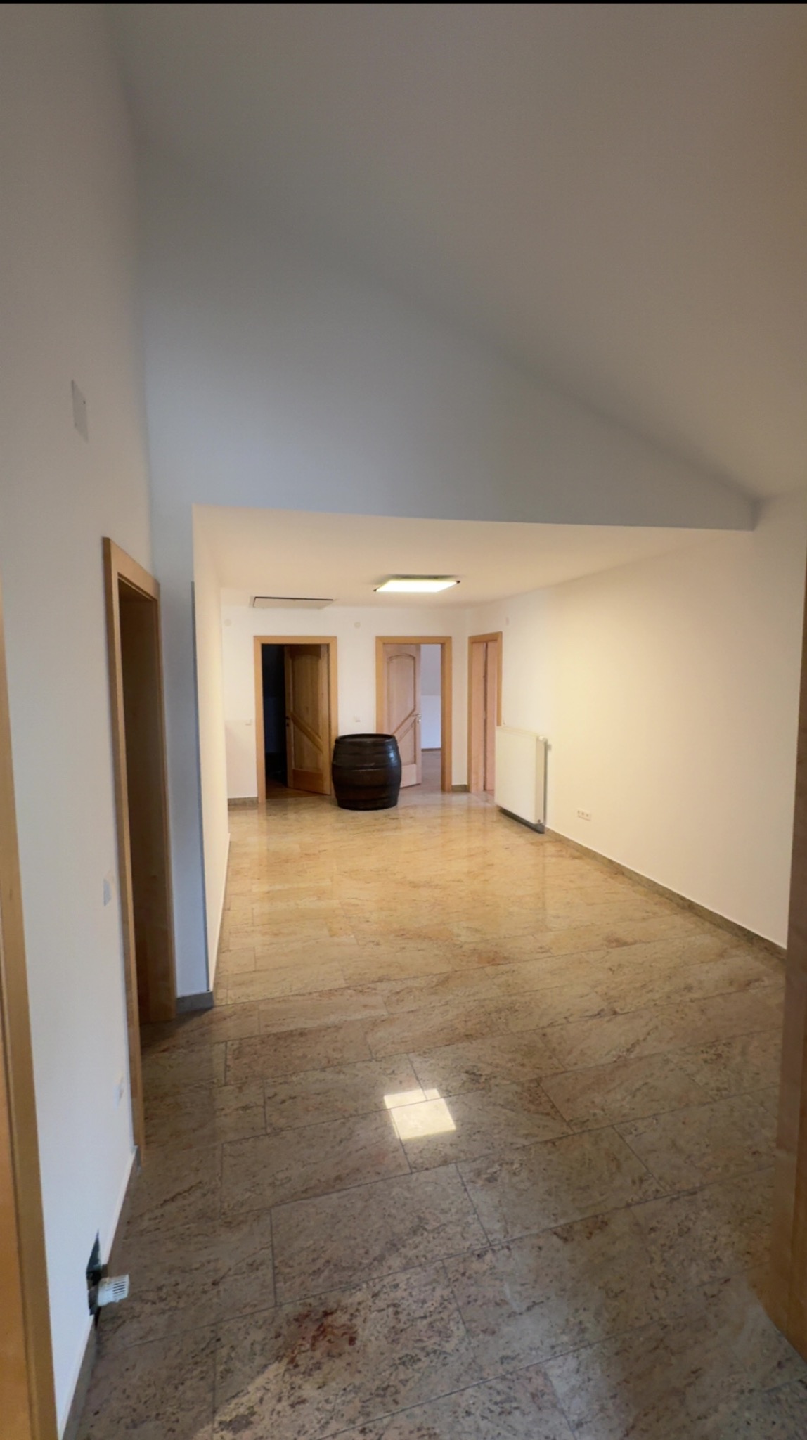 Przestronne wnętrze po remoncie z beżową podłogą z płytek, białymi ścianami i drewnianymi drzwiami. W tle widoczna beczka i grzejnik.