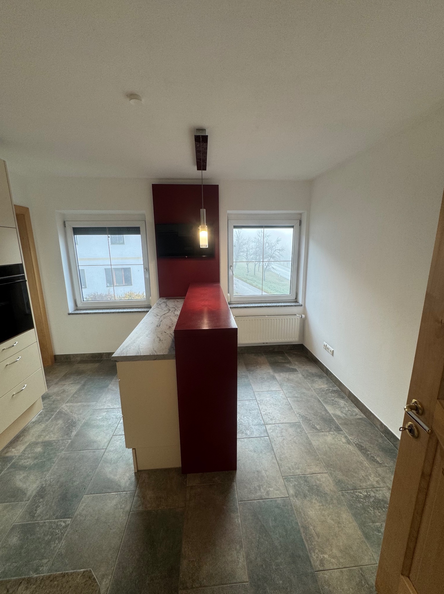 Nowoczesna kuchnia z wyspą i płytkami podłogowymi w odcieniach szarości. Czerwony panel ścienny z telewizorem. Widok z okna na zewnątrz. Minimalistyczny design.