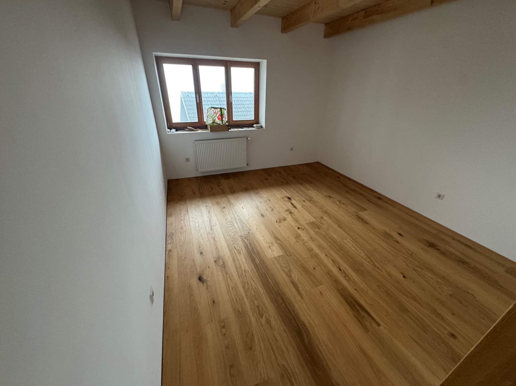 Jasne panele podłogowe z naturalnego drewna w pokoju z oknem i białymi ścianami. Widoczne drewniane belki stropowe. Minimalistyczny wystrój wnętrza.