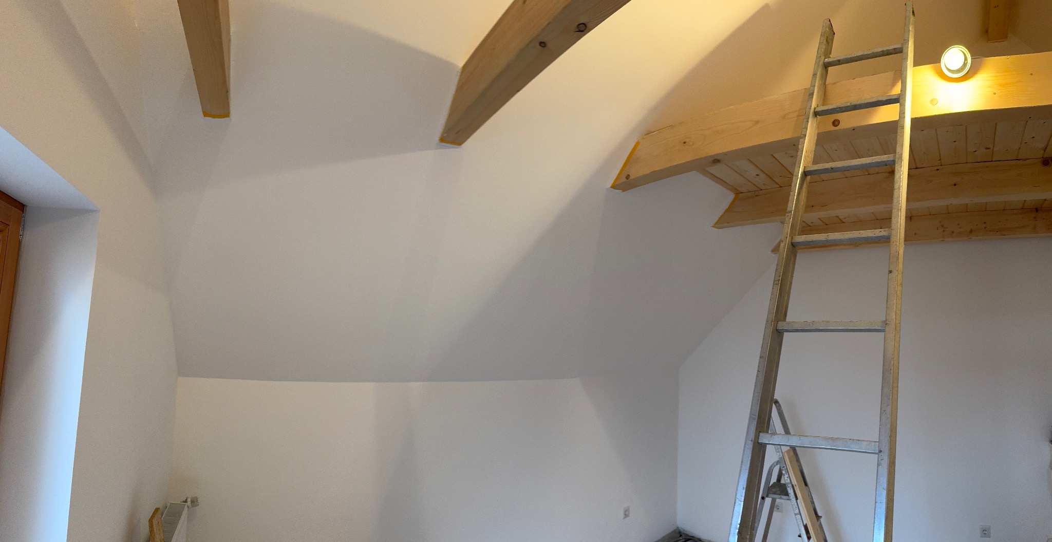 Białe poddasze po malowaniu z drewnianymi belkami i drabiną. Świeżo pomalowane ściany, widoczne zabezpieczenia taśmą malarską na belkach.