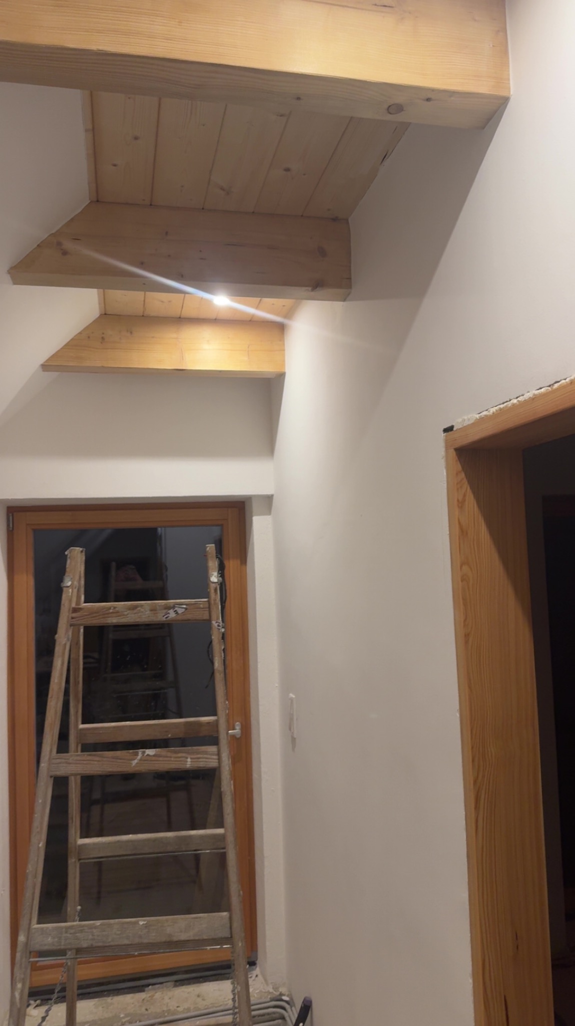 Drewniane belki stropowe w jasnym wnętrzu, widoczne z dołu. Drabina oparta o ścianę, okno w tle. Elementy wykończeniowe w trakcie prac.