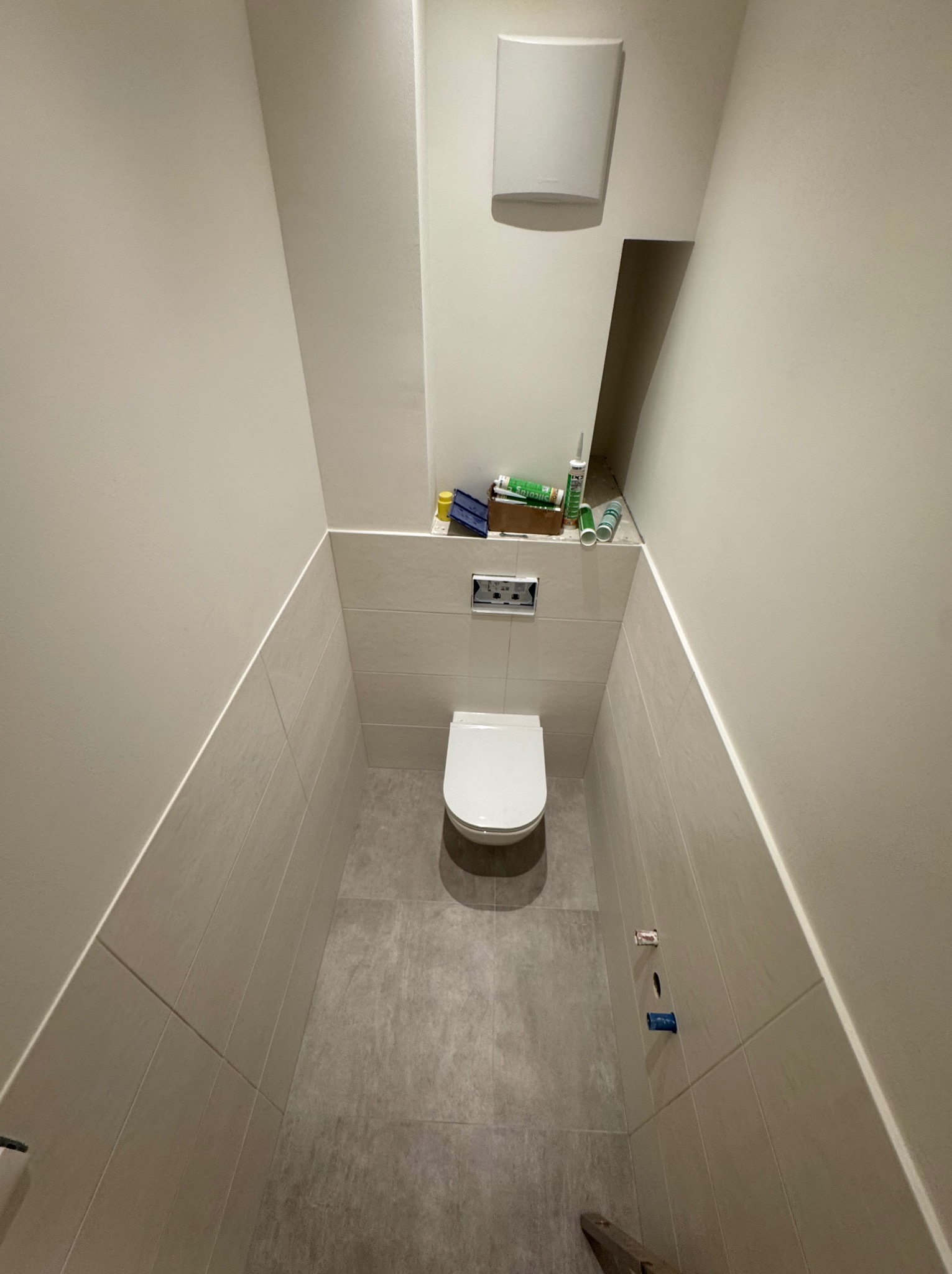 Wąska, wykafelkowana toaleta w odcieniach szarości i beżu. Podwieszana muszla klozetowa, zabudowa geberit, widoczne akcesoria montażowe. Minimalistyczny design.