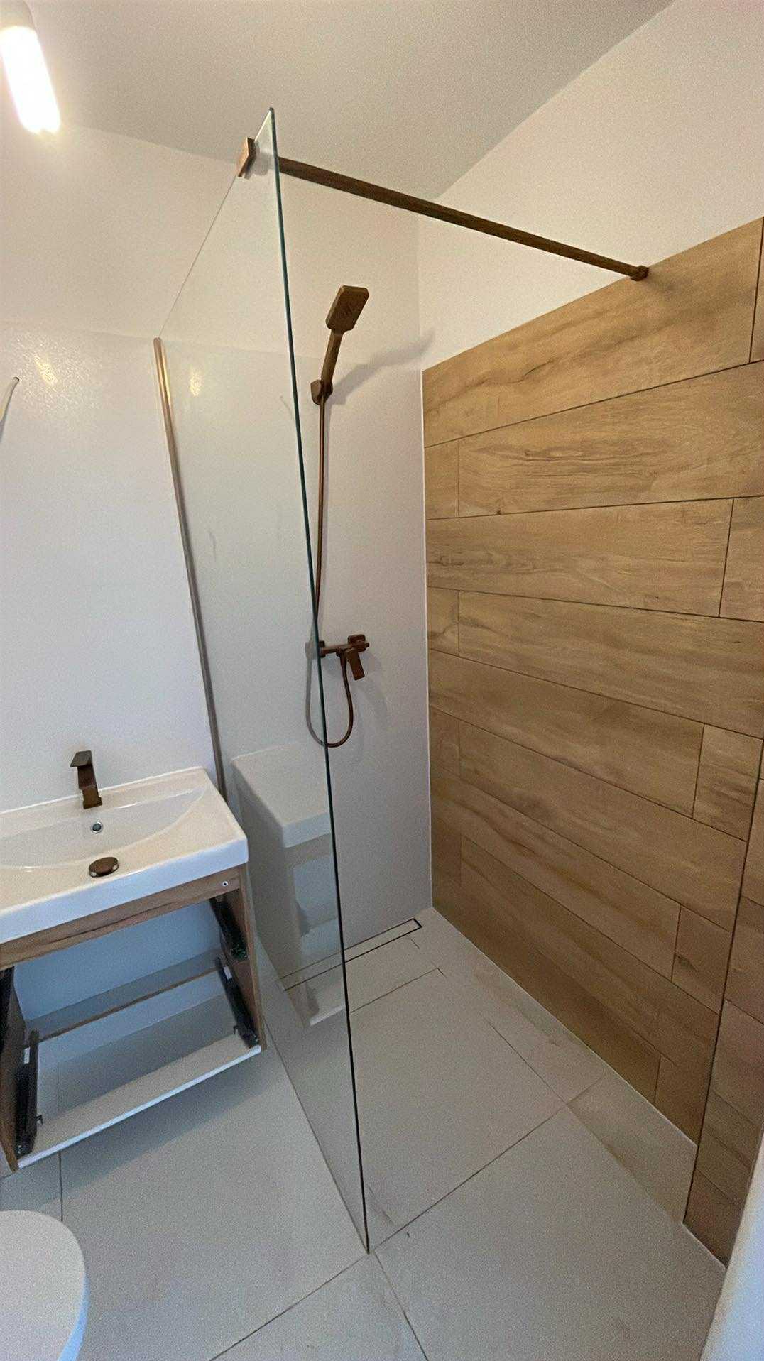 Nowoczesna łazienka z kabiną prysznicową typu walk-in, drewnopodobnymi płytkami na ścianie i umywalką z szafką. Armatura w kolorze miedzi.