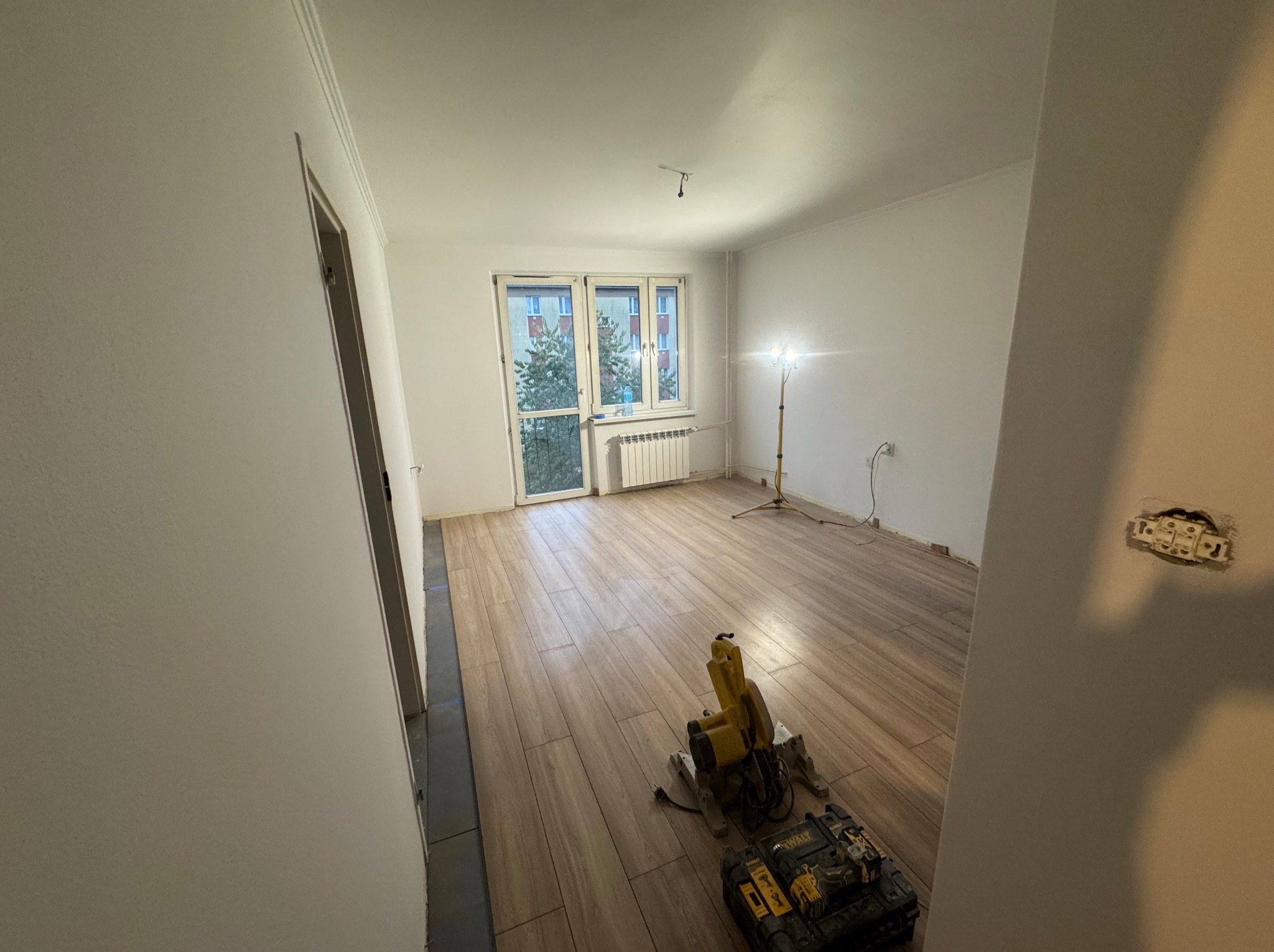 Układanie paneli podłogowych imitujących drewno w jasnym pokoju. Widoczna piła do cięcia paneli i lampa oświetlająca miejsce pracy. Częściowo wykończone pomieszczenie.