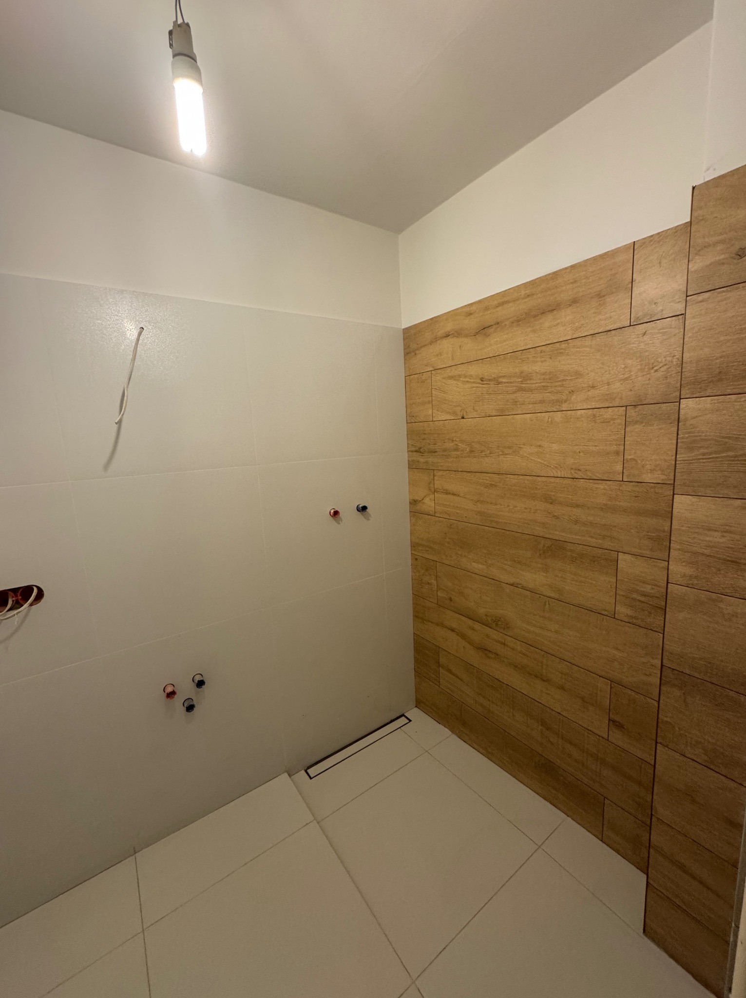 Wykończona łazienka z drewnopodobnymi i białymi płytkami, widoczne przyłącza wodne i odpływ liniowy. Oświetlenie sufitowe.