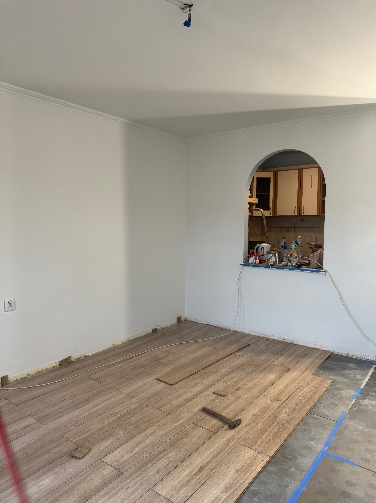 Układanie paneli podłogowych w jasnym wnętrzu z łukowym oknem na kuchnię. Częściowo ułożone panele imitujące drewno, widoczne narzędzia i fragmenty surowej podłogi.