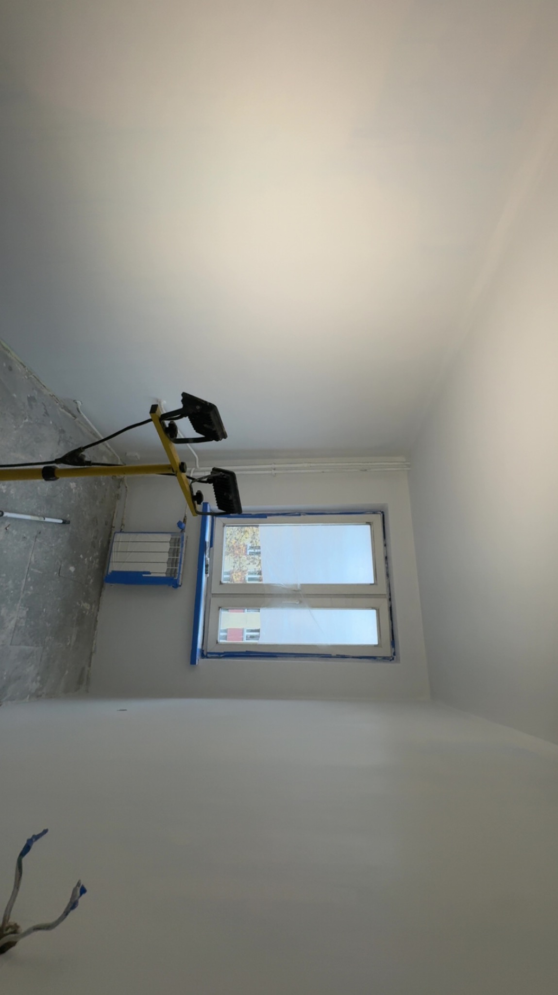 Wnętrze pokoju po malowaniu na biało, z widocznym oknem zabezpieczonym taśmą malarską i lampą halogenową na statywie. Świeżo pomalowane ściany i sufit.