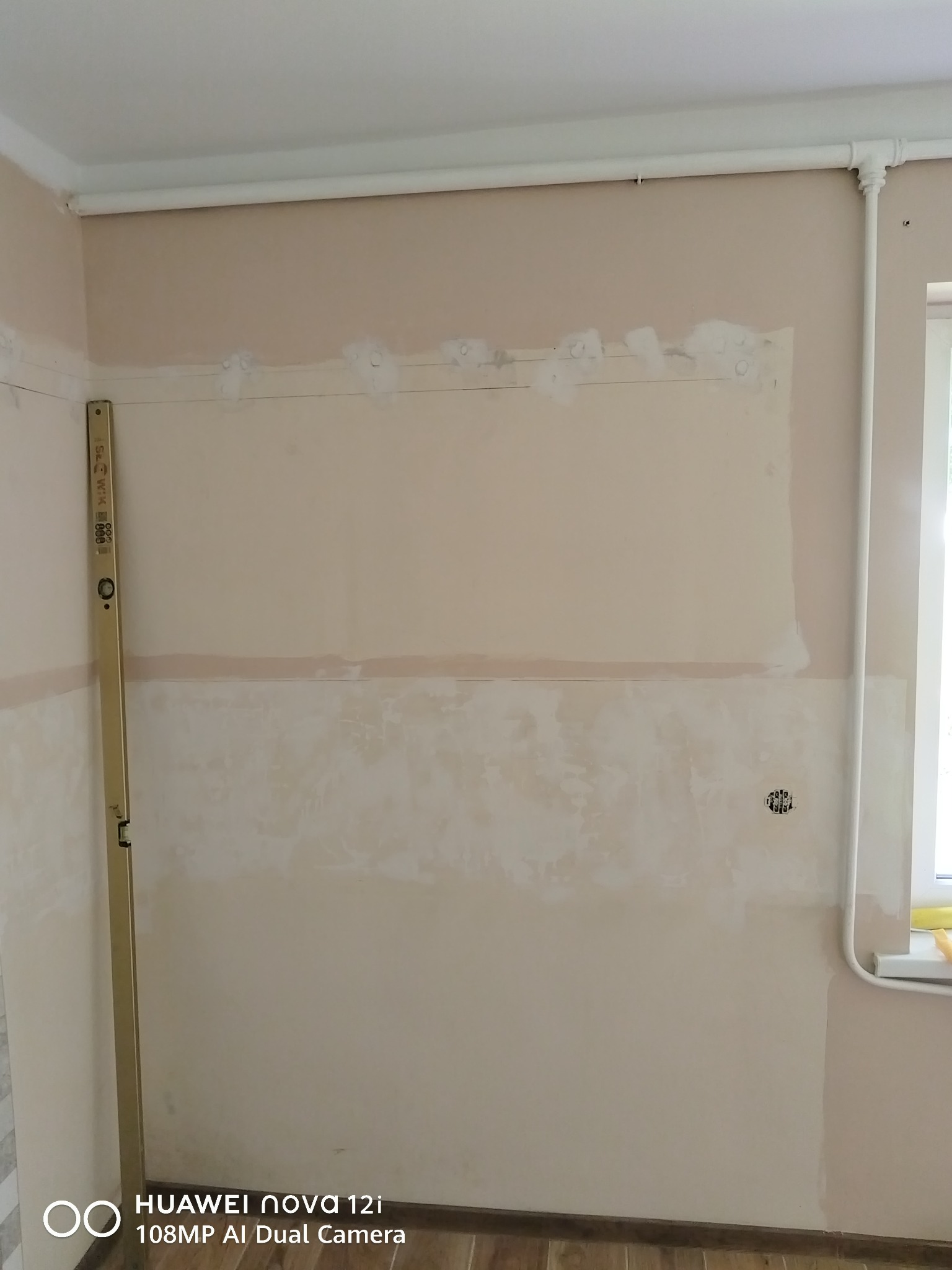 Ściana w trakcie przygotowania do malowania: szpachlowanie, gruntowanie. Widoczna poziomica przy ścianie i instalacja grzewcza. Etap prac wykończeniowych.