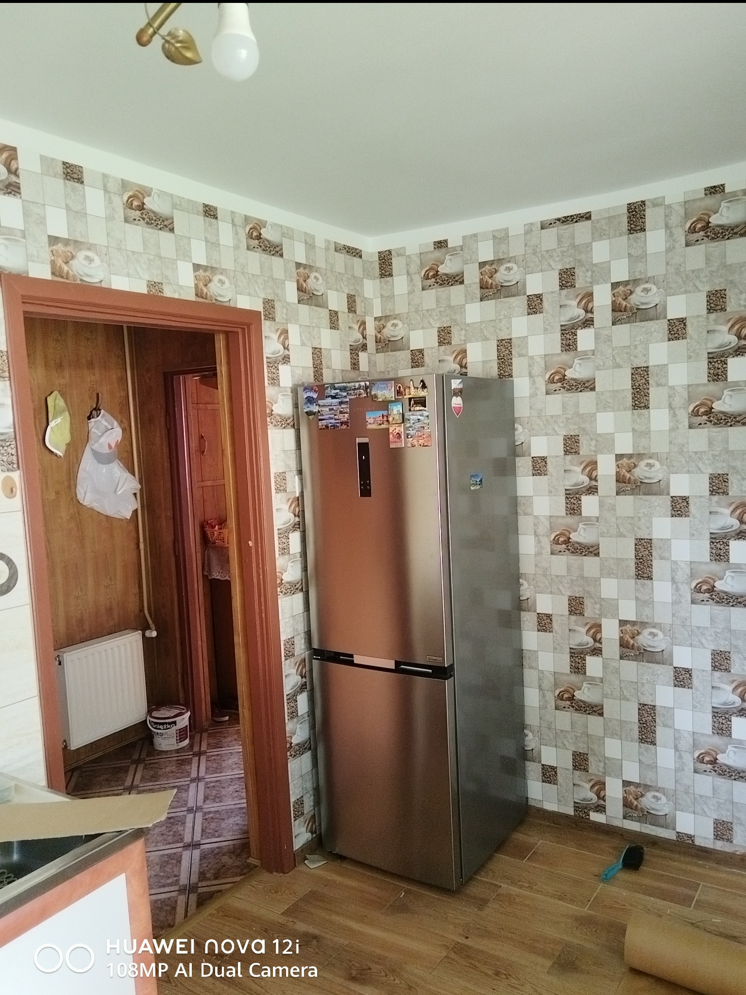 Kuchnia po remoncie w Szczepanowie: ściany w kwadratowe wzory z motywem kawy i rogalików, brązowe panele podłogowe, srebrna lodówka i widok na sąsiednie pomieszczenie.