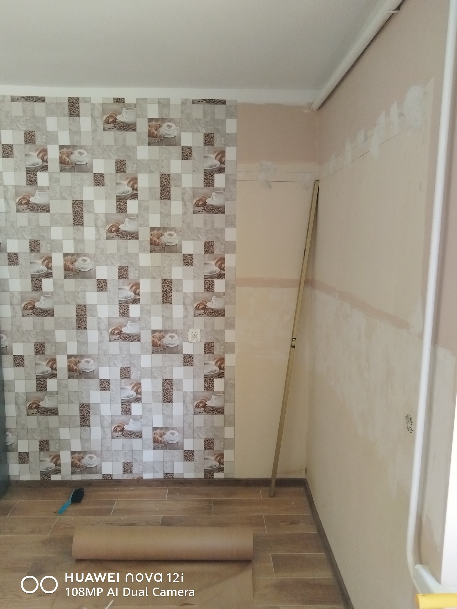 Fragment kuchni w trakcie tapetowania: wzorzysta tapeta z motywem kawy na jednej ścianie, surowa ściana obok, poziomica i rolka tapety na podłodze.