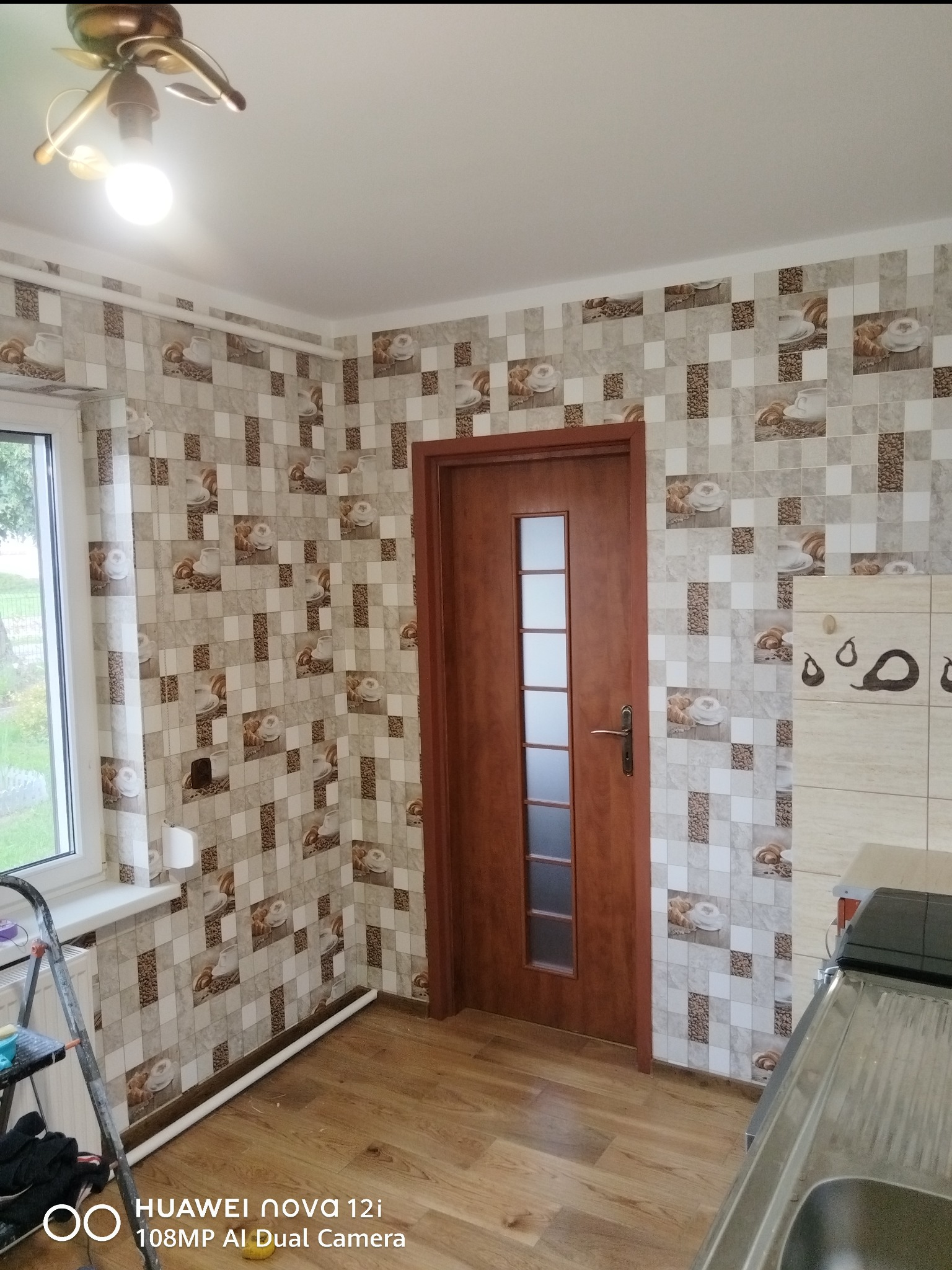 Wykończona kuchnia z tapetą w motywy kawowe i drewnianą podłogą. Widoczna brązowa ościeżnica drzwiowa i fragment zlewu. Ujęcie z dołu, widoczny fragment drabiny.