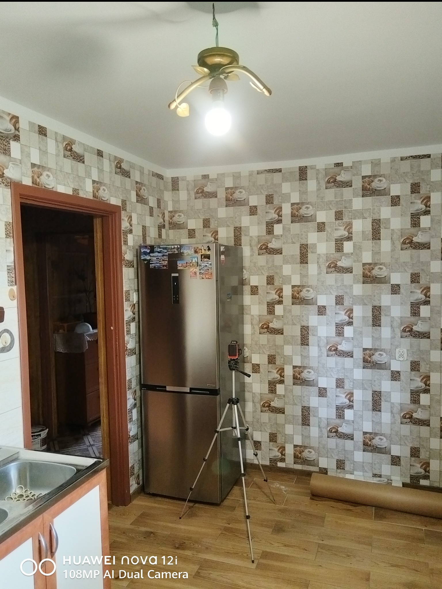 Kuchnia po tapetowaniu: ściany w mozaikę z motywem kawy i rogalików, srebrna lodówka, drewniana podłoga i statyw z telefonem. Widoczny fragment zlewu.
