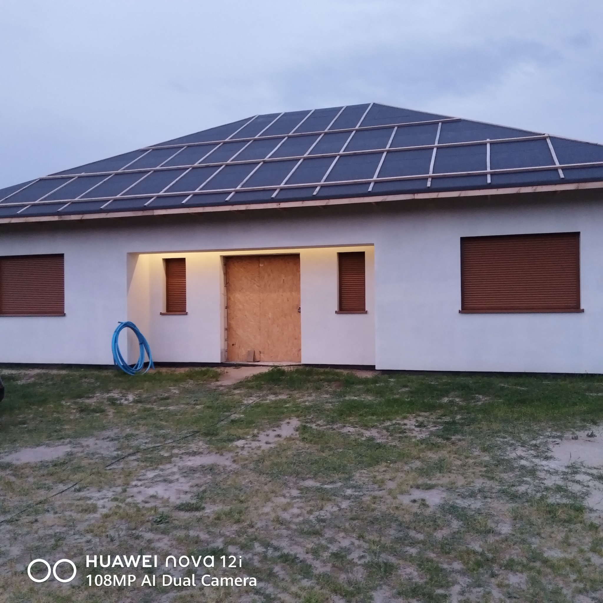 Biały parterowy dom z brązowymi roletami i przygotowanym dachem pod montaż paneli słonecznych. Wejście zabezpieczone płytą. Na trawniku zwinięty niebieski wąż.