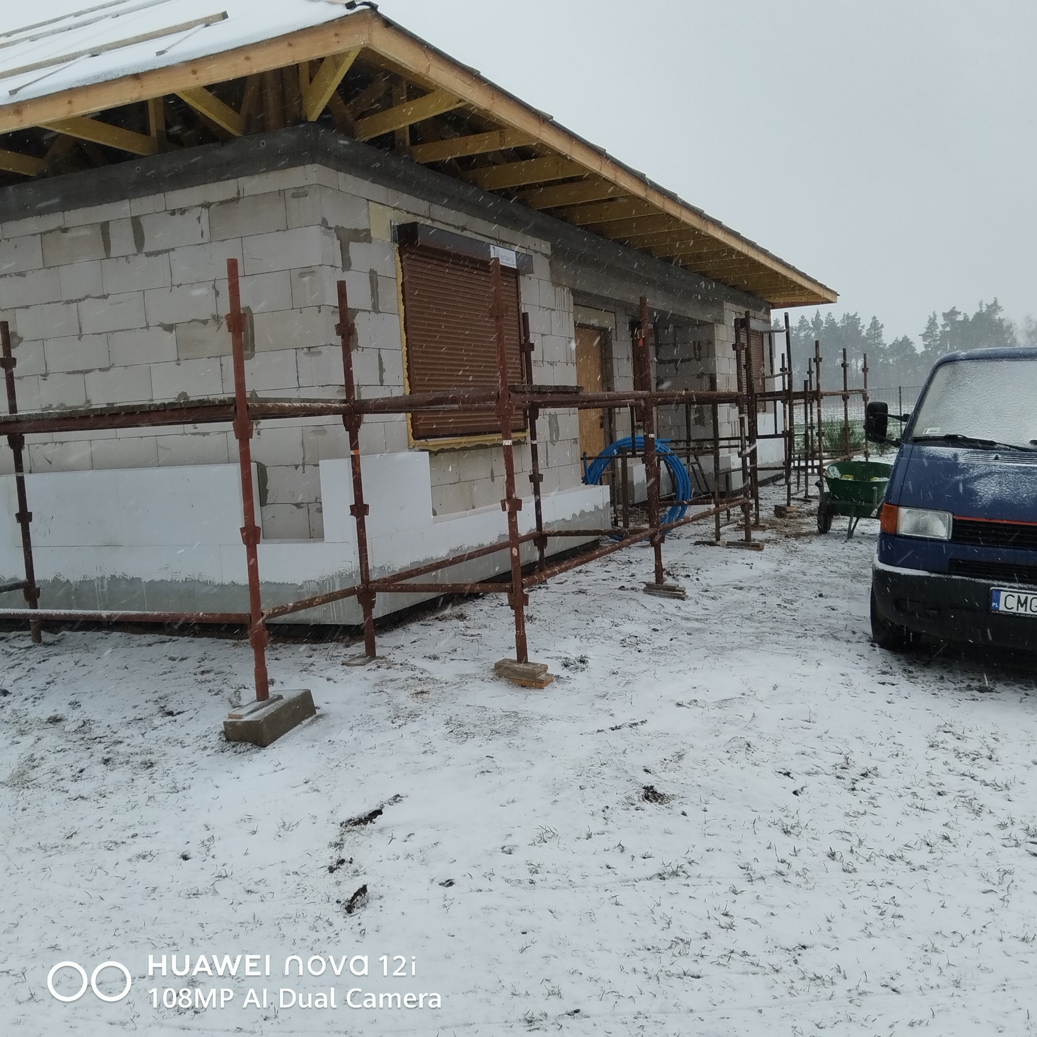 Budowa domu w zimowej scenerii: ściany z bloczków, drewniana konstrukcja dachu, rusztowania i częściowo ocieplona elewacja styropianem. Na pierwszym planie ośnieżony teren.