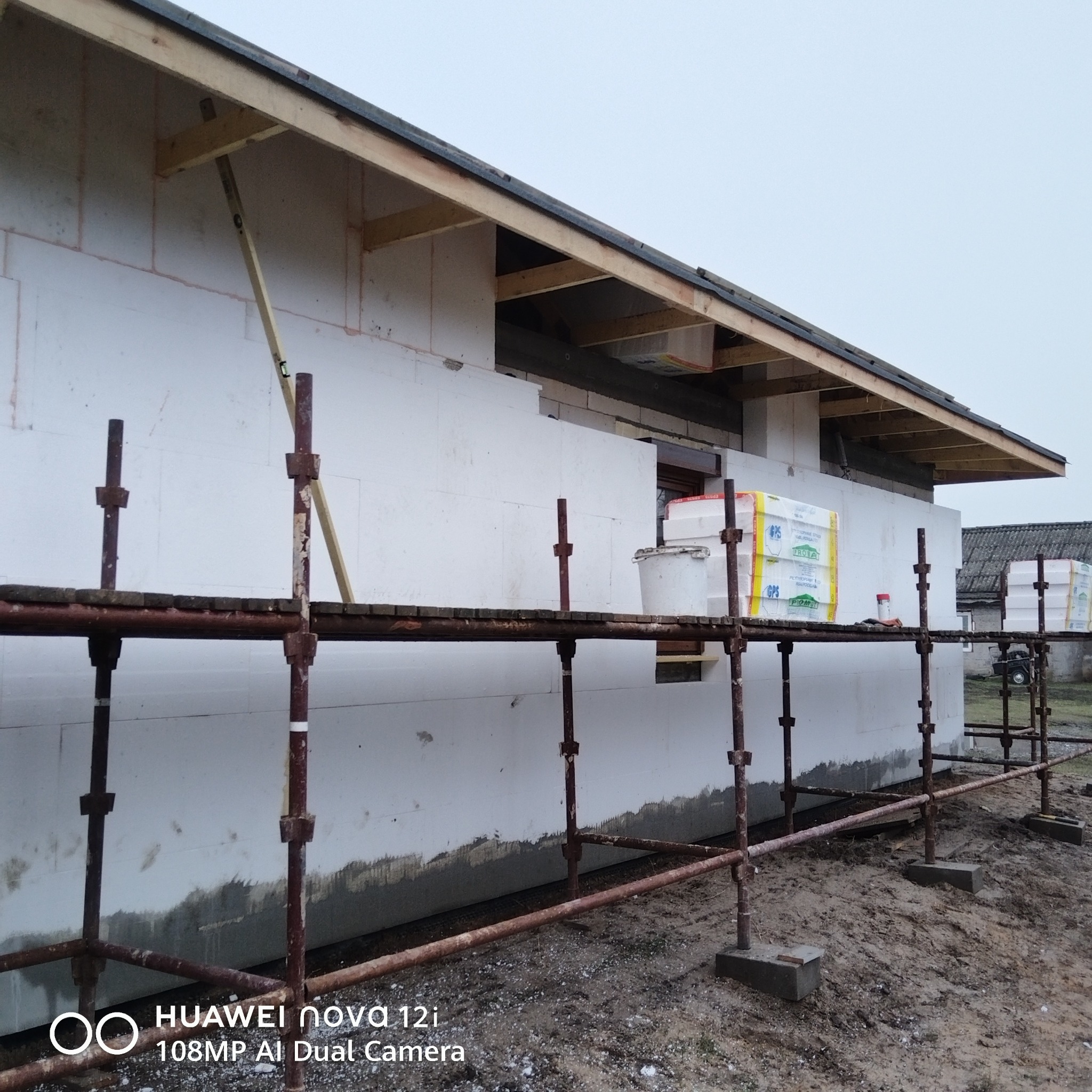 Budowa domu w trakcie ocieplania. Widoczne rusztowania, białe płyty izolacyjne na ścianach i drewniana konstrukcja dachu. Na rusztowaniu paczki materiałów izolacyjnych.