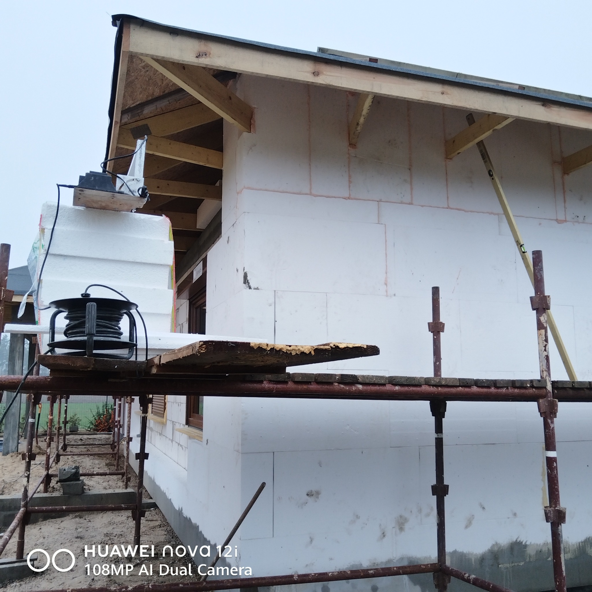 Ocieplanie budynku styropianem na rusztowaniu. Widoczne białe płyty izolacyjne i drewniana konstrukcja dachu. Prace w toku, surowy stan.