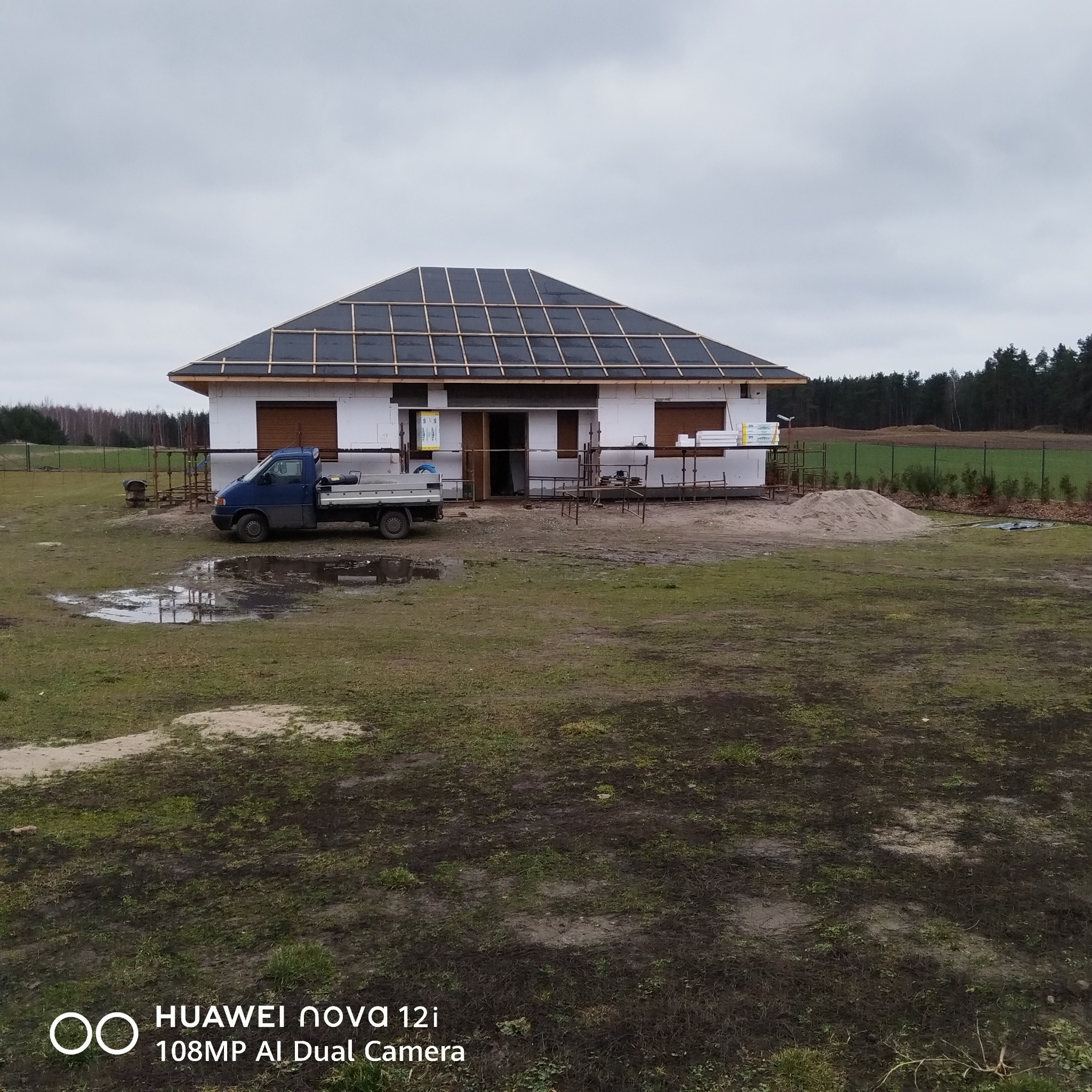 Dom w trakcie ocieplania elewacji, widoczne płyty izolacyjne i konstrukcja dachu. Na pierwszym planie zaparkowany samochód dostawczy.