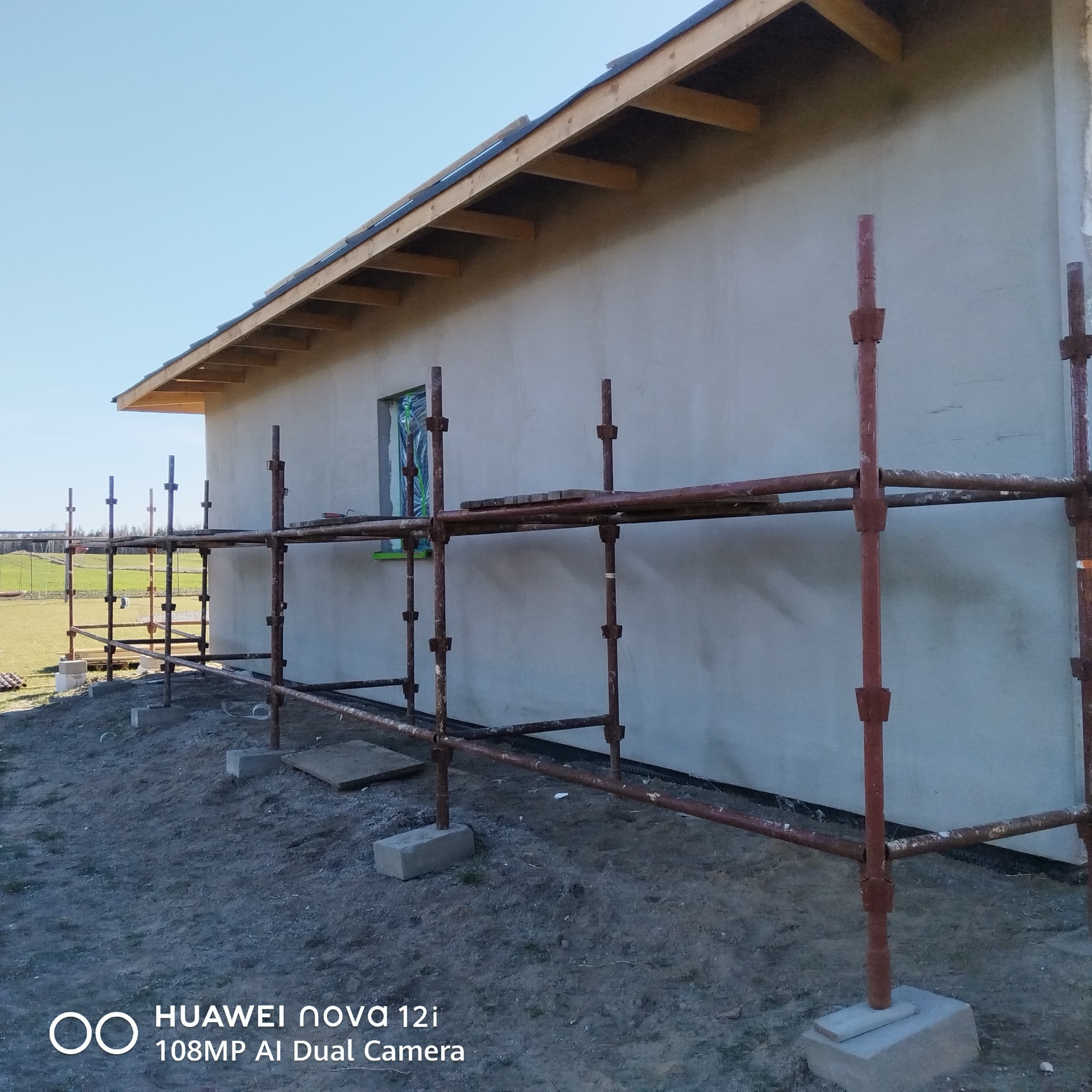 Ściana budynku w trakcie ocieplania, widoczne rusztowania i okno zabezpieczone folią. Surowy stan elewacji, przygotowanie do dalszych prac wykończeniowych.