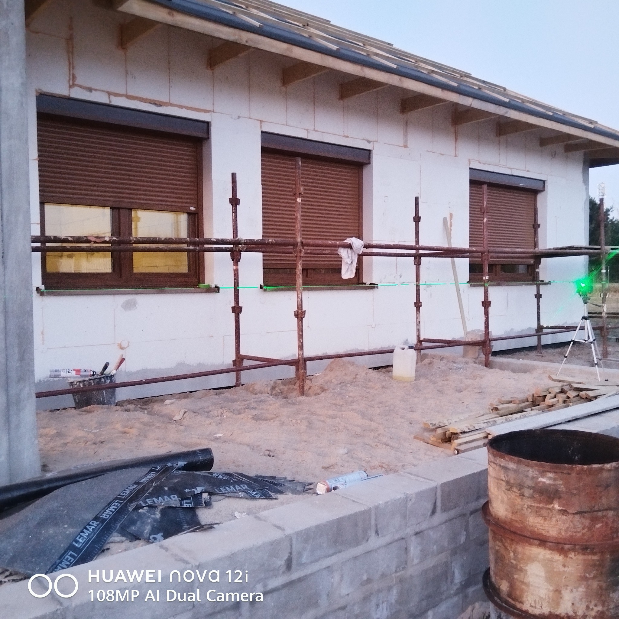 Budynek w trakcie ocieplania styropianem, widoczne rusztowania, okna z roletami, poziomica laserowa i narzędzia. Etap prac przed wykończeniem elewacji.