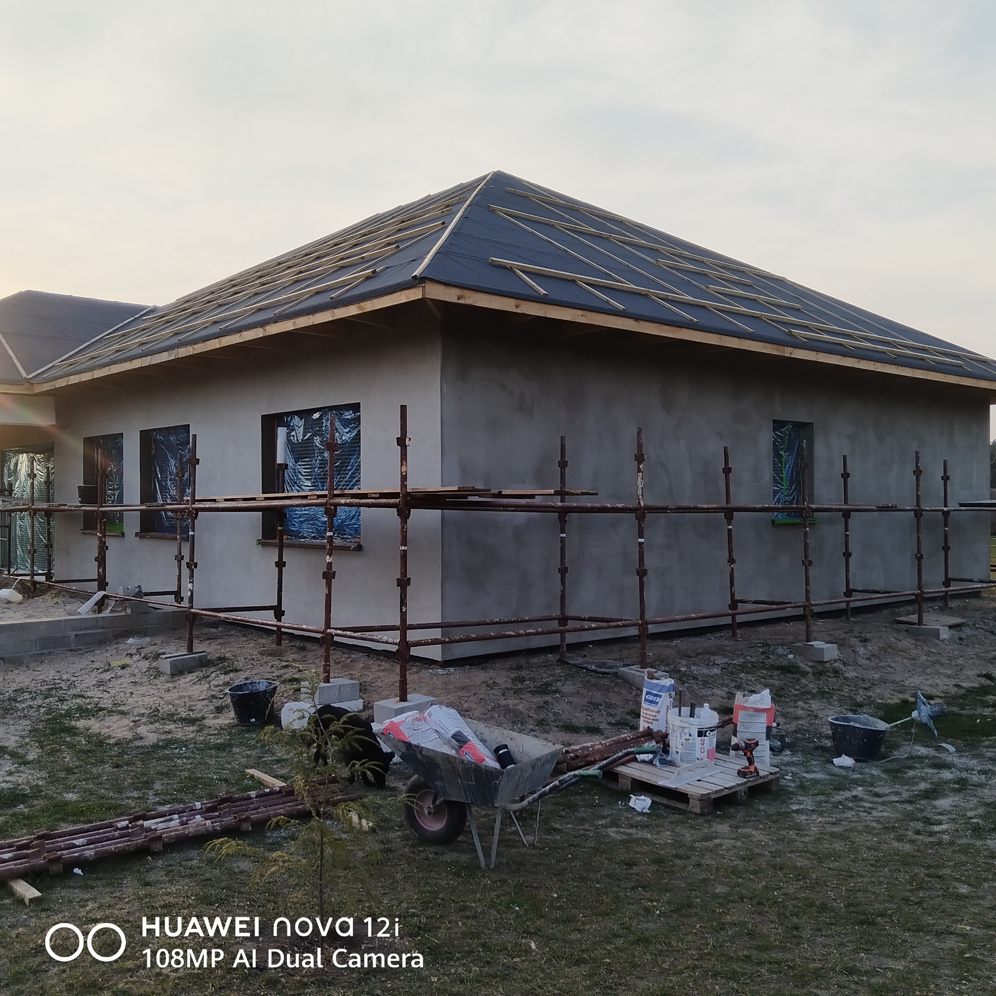 Surowy dom jednorodzinny w trakcie ocieplania elewacji, z widocznymi rusztowaniami i materiałami budowlanymi. Dach pokryty papą, okna zabezpieczone folią.