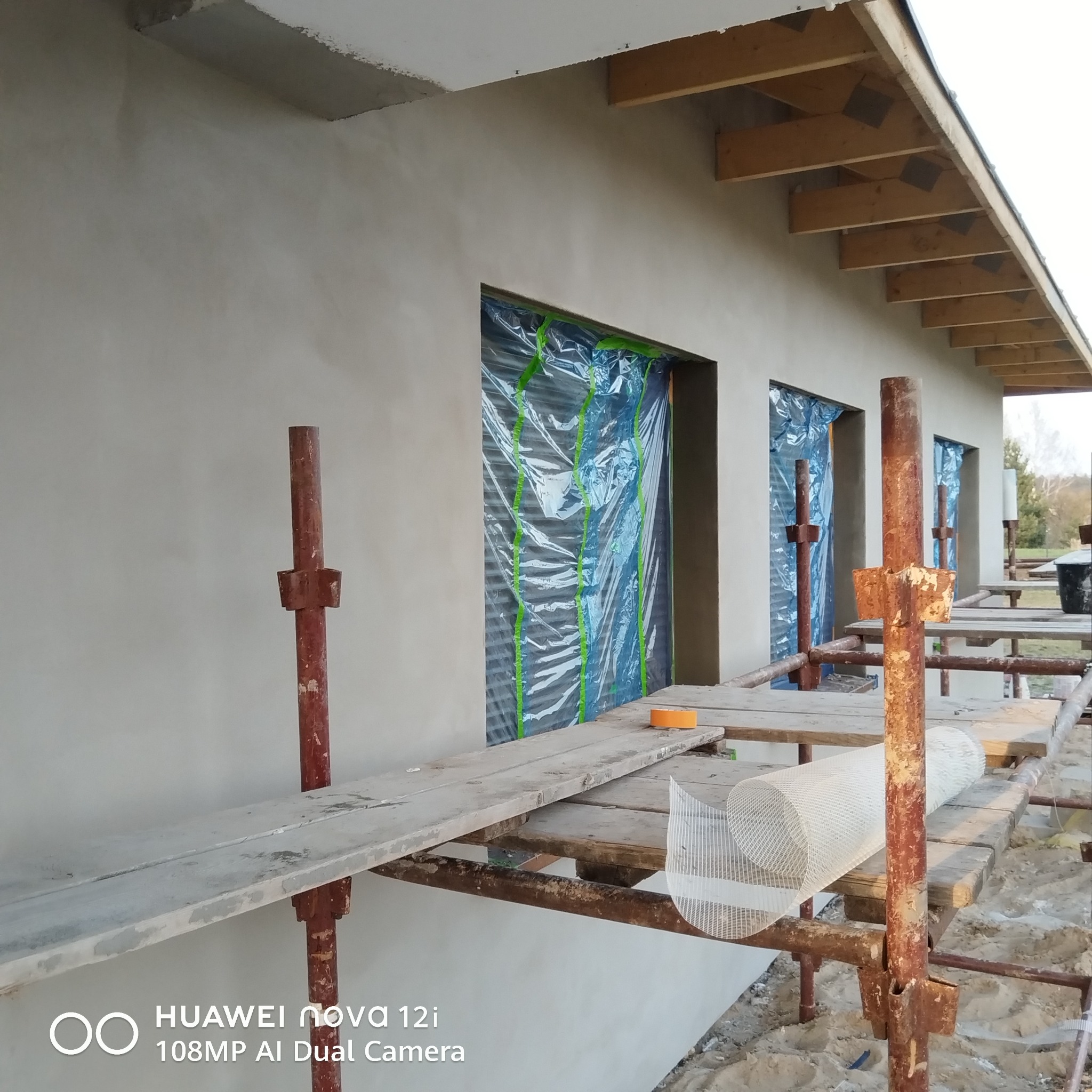 Ściana budynku po nałożeniu szarego tynku, okna zabezpieczone folią. Widoczne rusztowanie z materiałami budowlanymi. Prace wykończeniowe elewacji w toku.