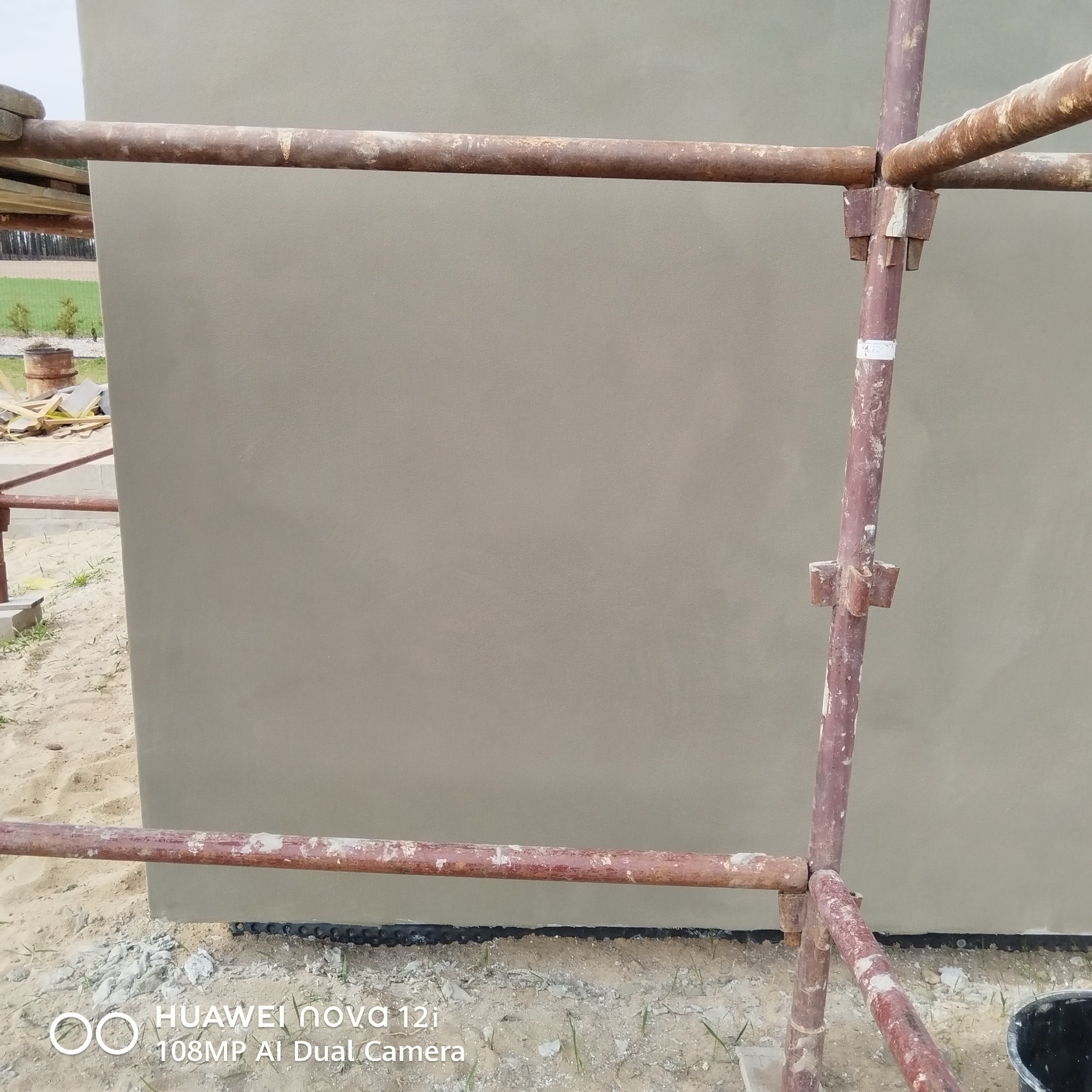 Świeżo otynkowana ściana zewnętrzna w kolorze beżowym, widoczna z bliska, z fragmentem rusztowania po bokach. Tło stanowi piaszczysta ziemia i fragment zieleni.