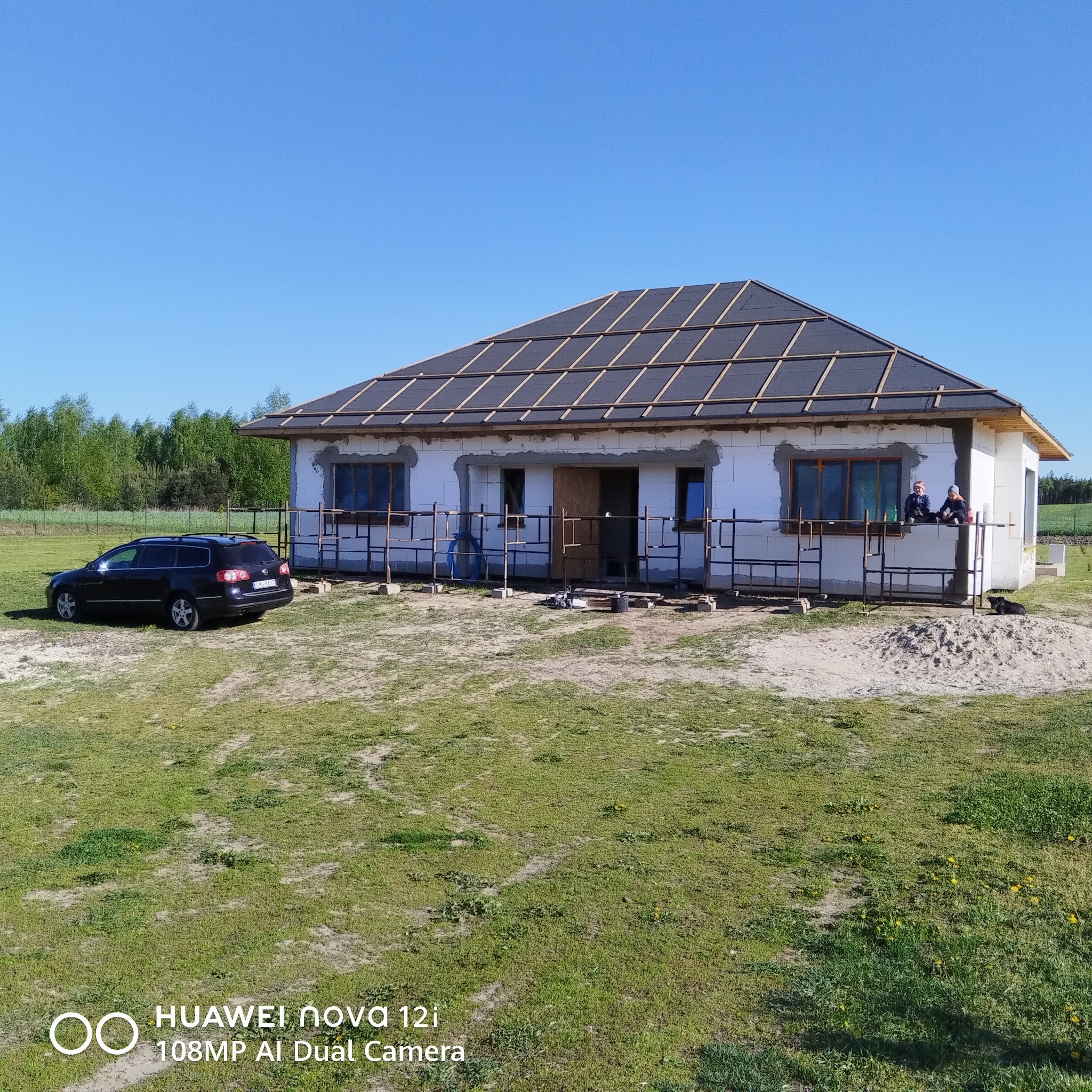 Dom w trakcie budowy z częściowo wykończonym dachem, widoczne rusztowania i samochód osobowy na trawniku. Dwóch pracowników siedzi na rusztowaniu.