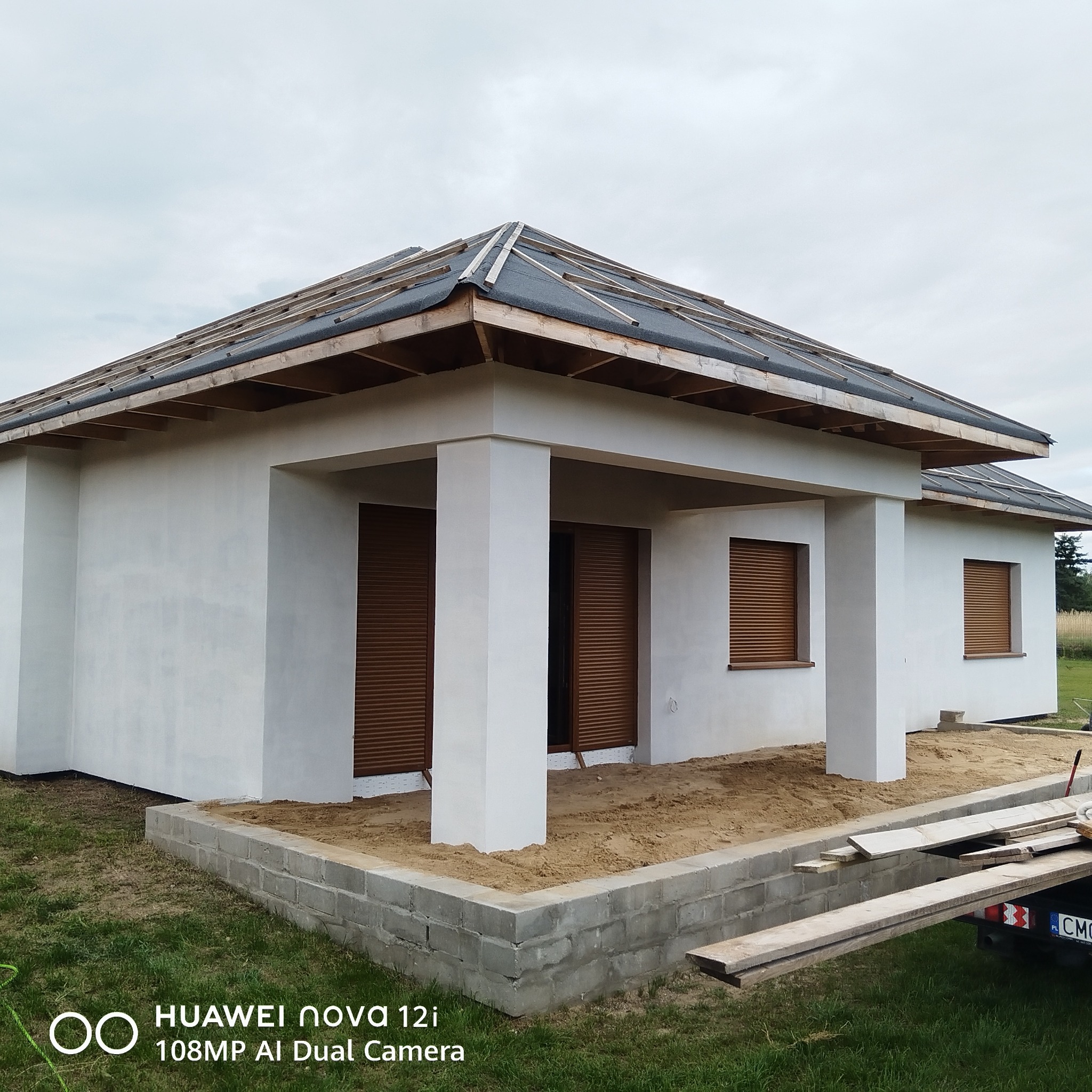 Nowoczesny dom jednorodzinny z białymi ścianami i brązowymi okiennicami w trakcie budowy dachu. Widoczne elementy konstrukcyjne dachu i przygotowany teren wokół budynku.