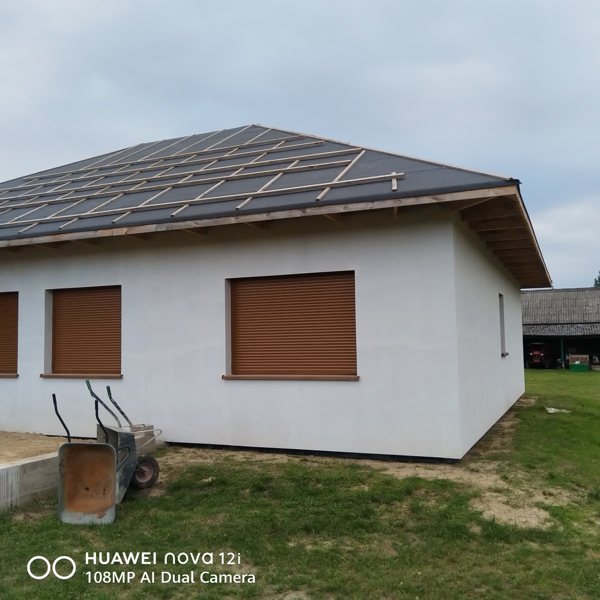 Biały dom z brązowymi roletami i przygotowanym dachem pod pokrycie. Dwie taczki stoją przed budynkiem na trawie. Widoczny etap budowy.