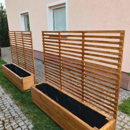 GranWood - Dwie drewniane donice z kratownicą, wyłożone czarną folią, stoją na trawie obok chodnika z kostki. Donice są wykonane z jasnego drewna i mają prostą, nowoczesną formę.