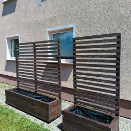 GranWood - Para ciemnobrązowych, drewnianych donic z wysokimi, lamelowymi kratkami, ustawionych na bruku obok trawnika przy budynku, z widocznymi czarnymi wkładami.