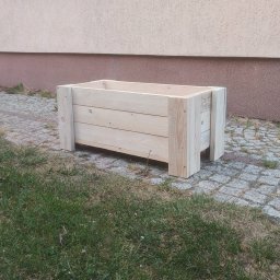 GranWood - Prostokątna donica z jasnego, surowego drewna, zbudowana z poziomych desek i pionowych wzmocnień, stojąca na bruku przy trawniku, na tle beżowej elewacji budynku.