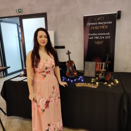 Skrzypcowe Historie - Kobieta w kwiecistej sukni prezentuje skrzypce i ofertę oprawy muzycznej na czarnym stole, oświetlone dekoracyjnymi lampkami. W tle baner z napisem 'Oprawa muzyczna SKRZYPCE'.