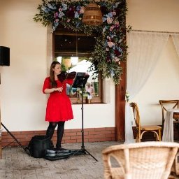 Skrzypcowe Historie - Skrzypaczka w czerwonej sukience gra na skrzypcach na tle dekoracji kwiatowych i białych zasłon, tworząc elegancką oprawę muzyczną wydarzenia.