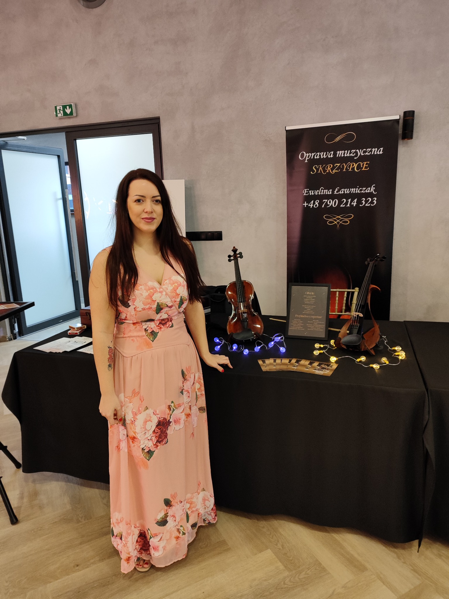 Kobieta w kwiecistej sukni prezentuje skrzypce i ofertę oprawy muzycznej na czarnym stole, oświetlone dekoracyjnymi lampkami. W tle baner z napisem 'Oprawa muzyczna SKRZYPCE'.